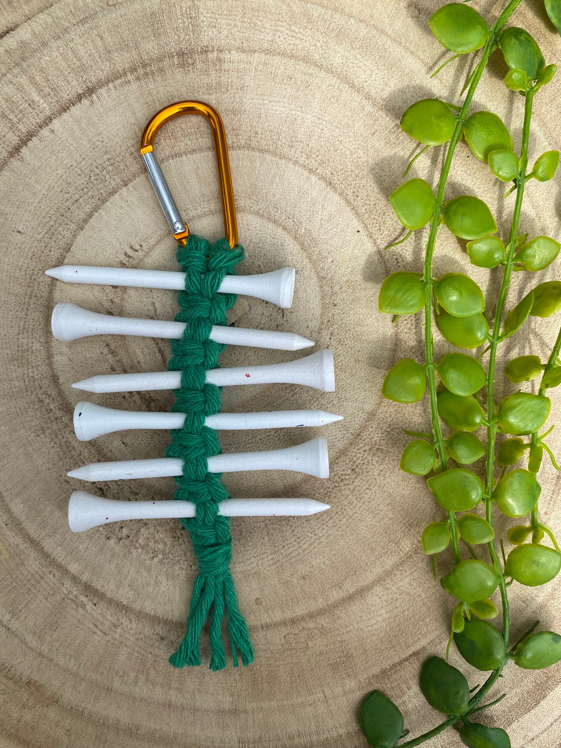 A knotted golf tee holder, made with green yarn on an orange gold carabiner clip, laid on a wooden surface holding 6 white golf tees.