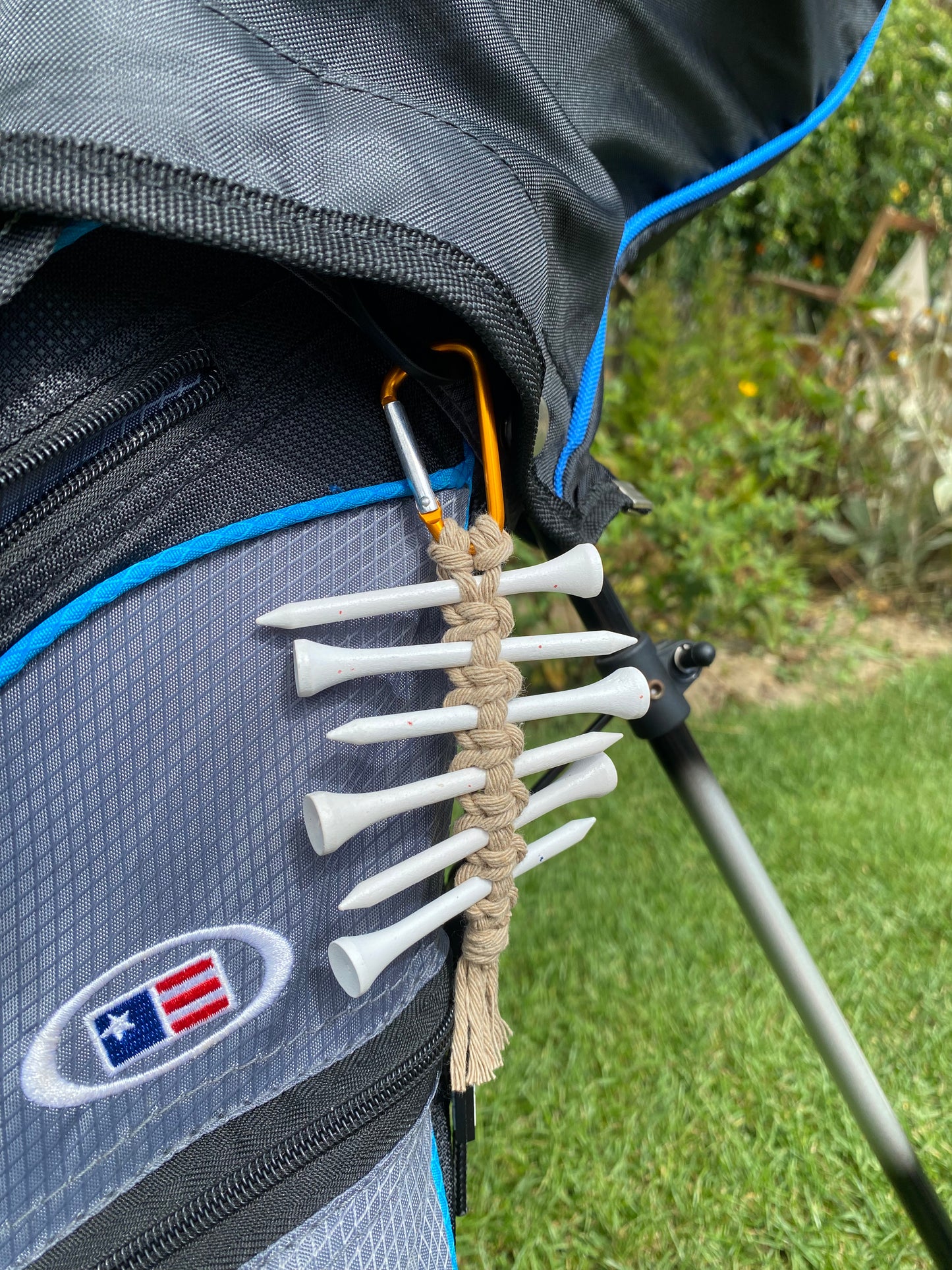 A knotted golf tee holder, made with beige yarn on an orange gold carabiner clip, attached to the side of a black golf bag, holding 6 white golf tees.