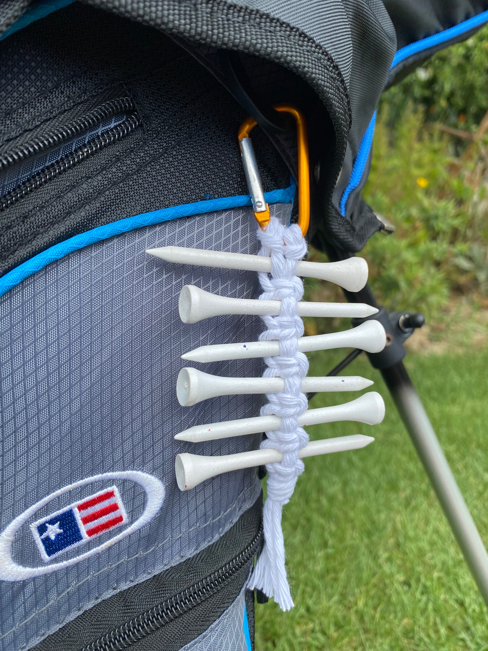 A knotted golf tee holder, made with white yarn on an orange gold carabiner clip, attached to a black golf bag, holding 6 white golf tees.