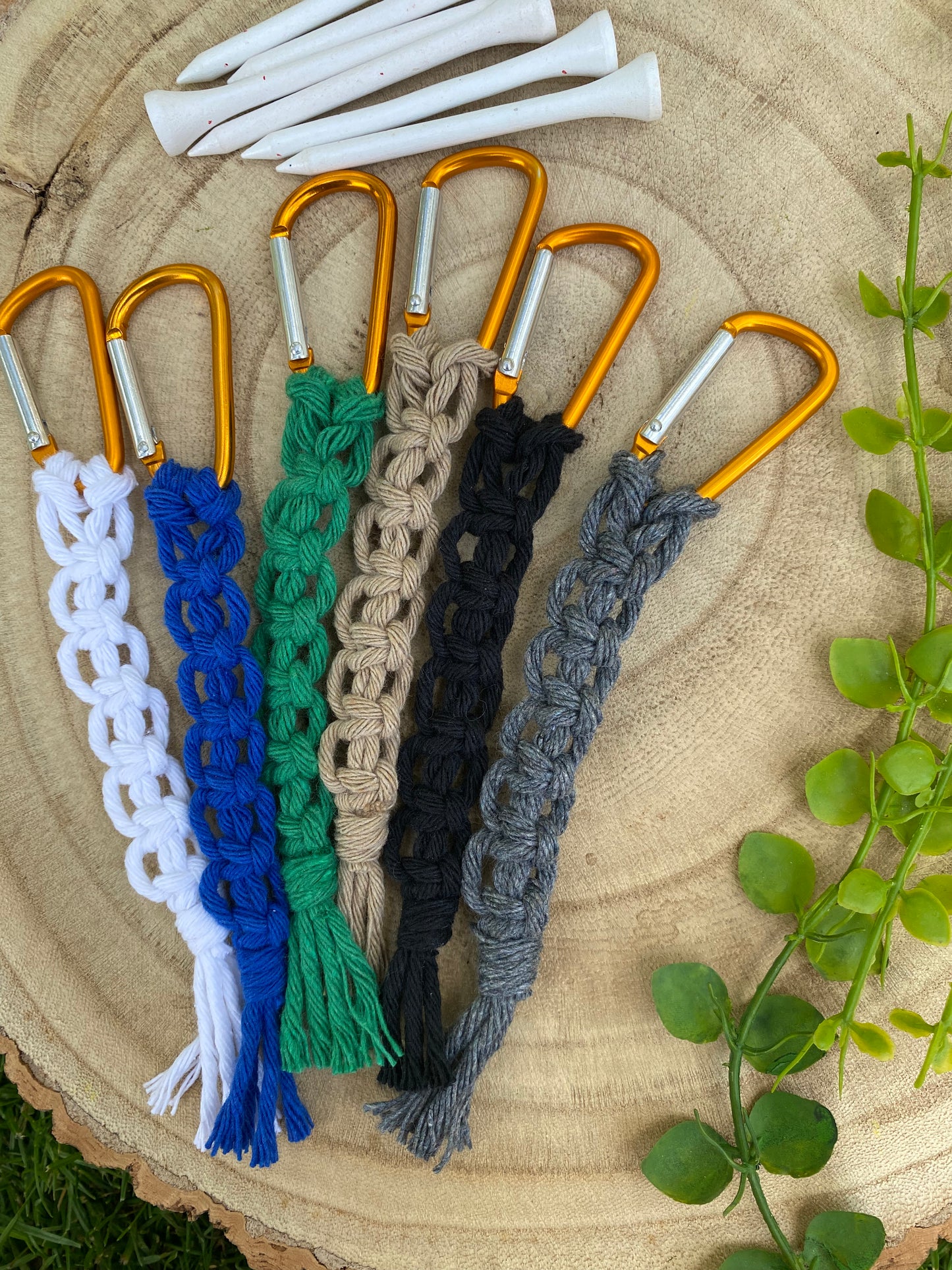 6 knotted golf tee holders, made with different coloured yarns on an orange gold carabiner clip, laid on a wooden surface next to 6 white golf tees.