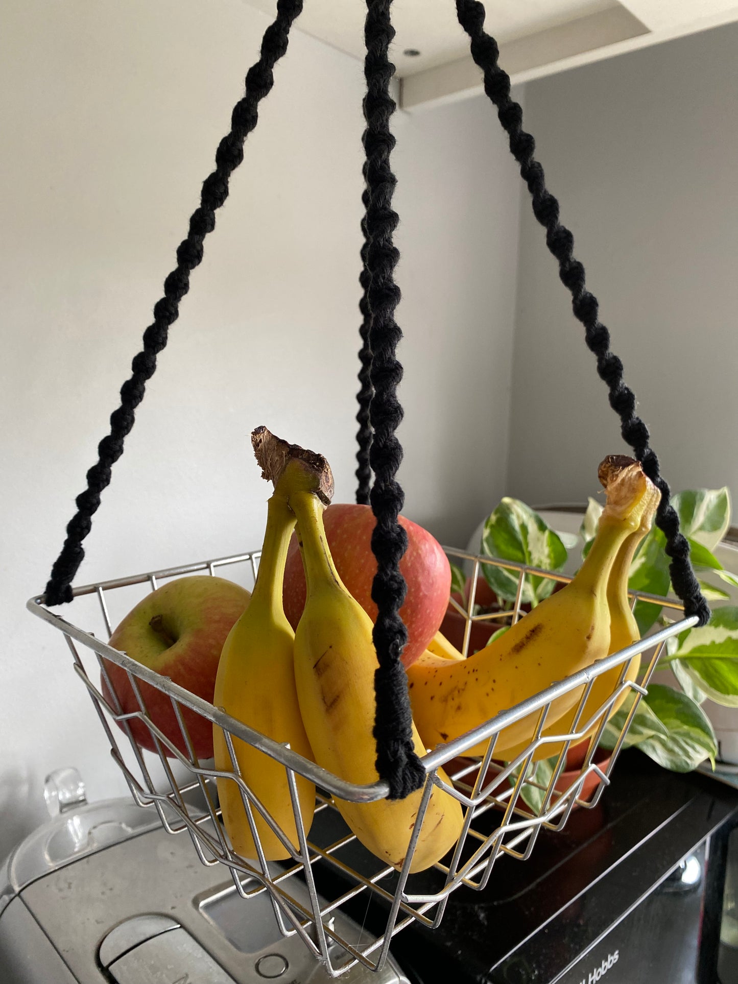 Close up of bananas and apples in a metal basket, suspended by black macrame ropes. There is a microwave and coffee machine underneath and a plant in the background.