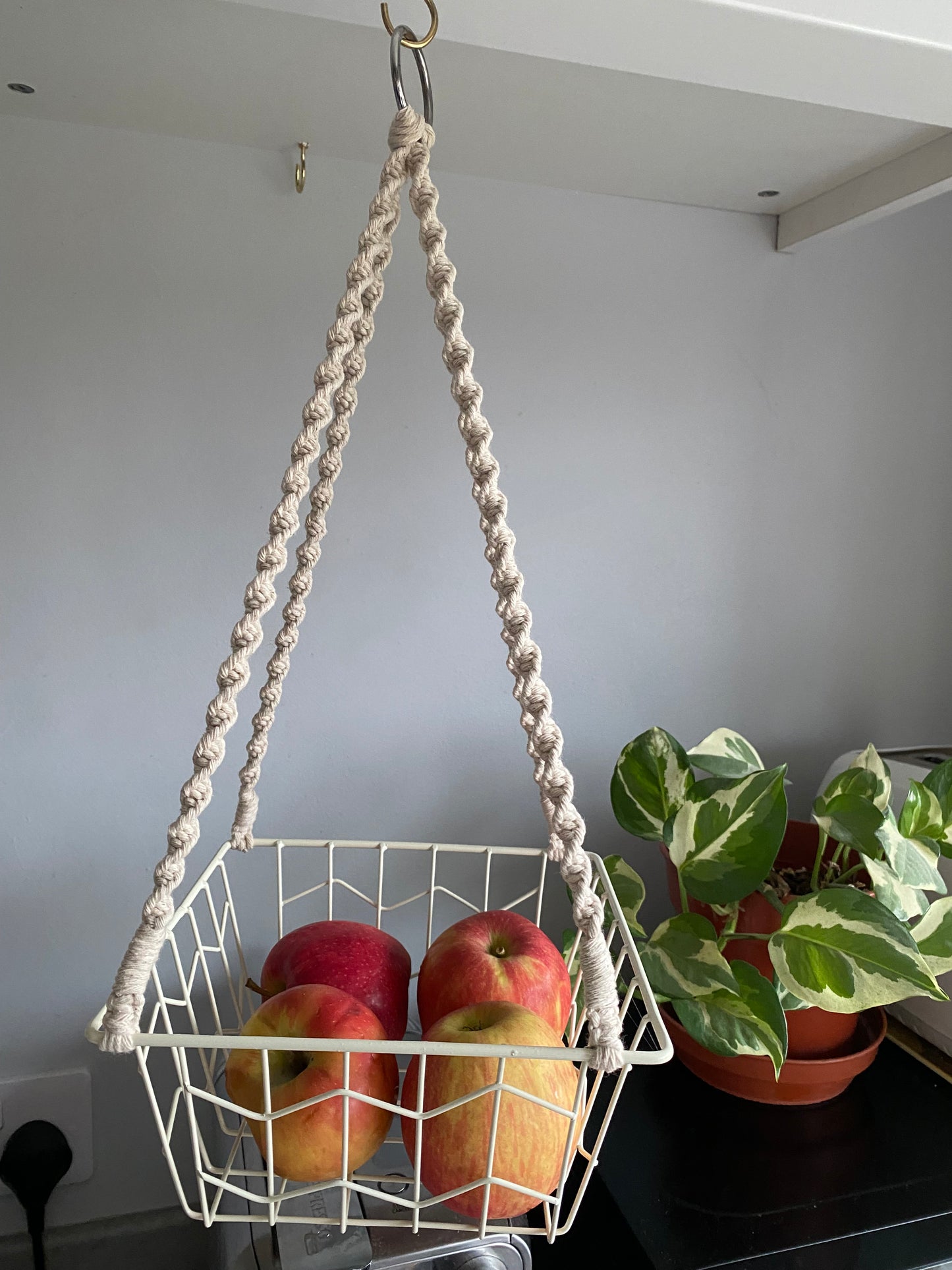 Four small red apples in a metal basket, suspended by natural macrame ropes on a metal hoop, hanging under a white kitchen cabinet next to a variegated houseplant.