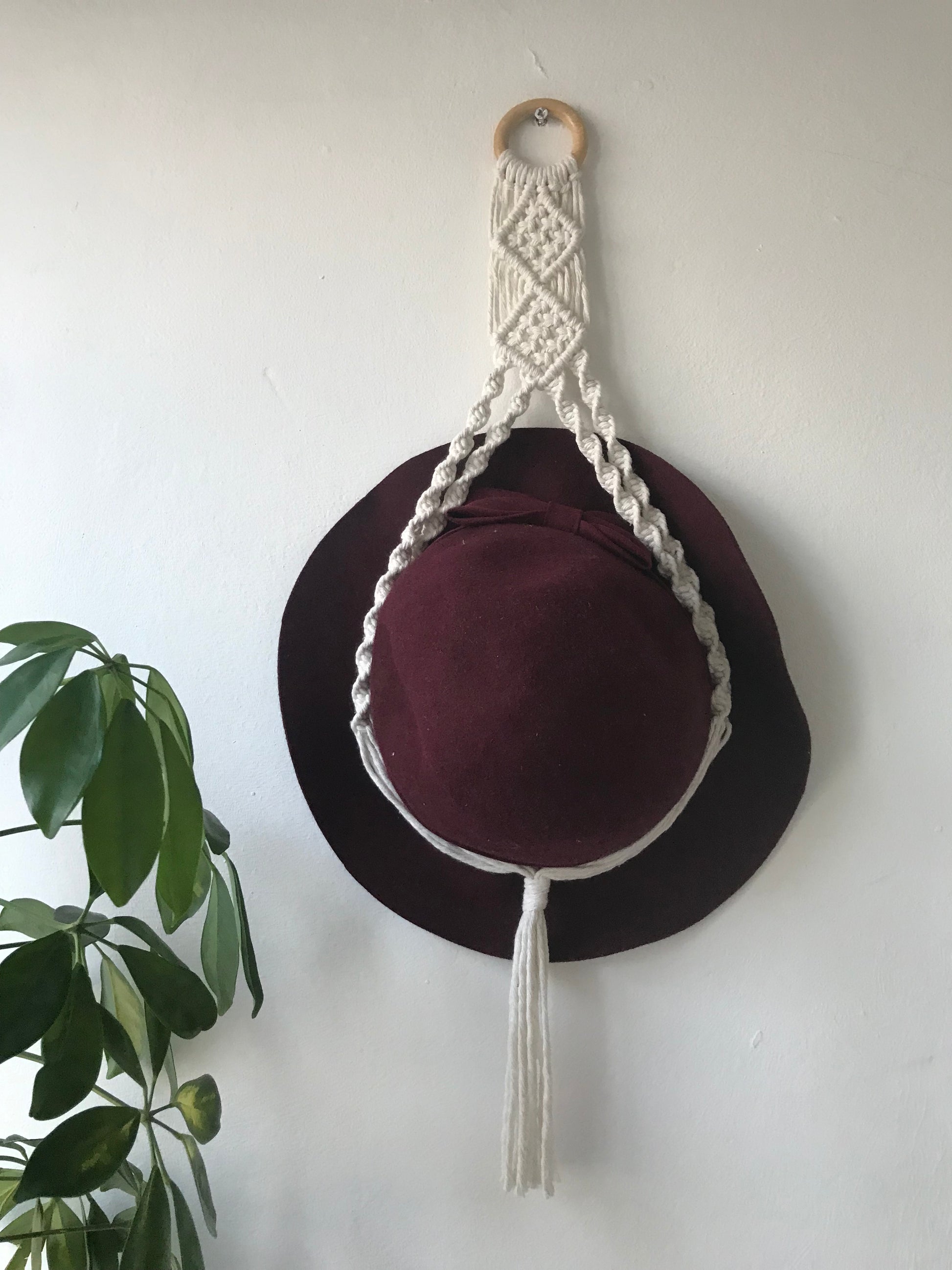 Macrame wall hanging hat display made with recycled cotton yarn in a natural colour. Shown with a dark red hat displayed inside it.