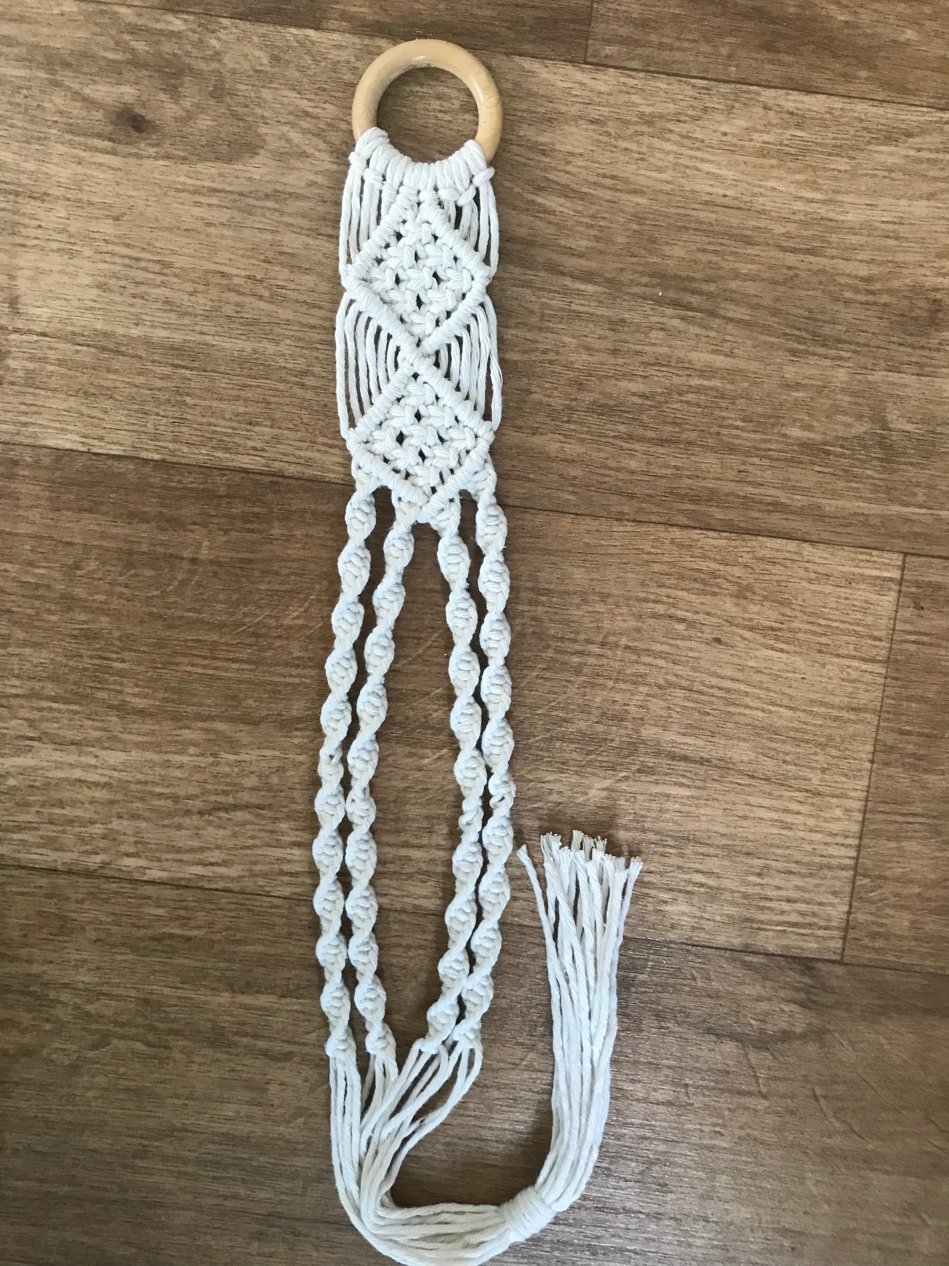 An empty macrame hat holder, made with natural coloured yarn and a wooden ring, laid on a wooden floor.