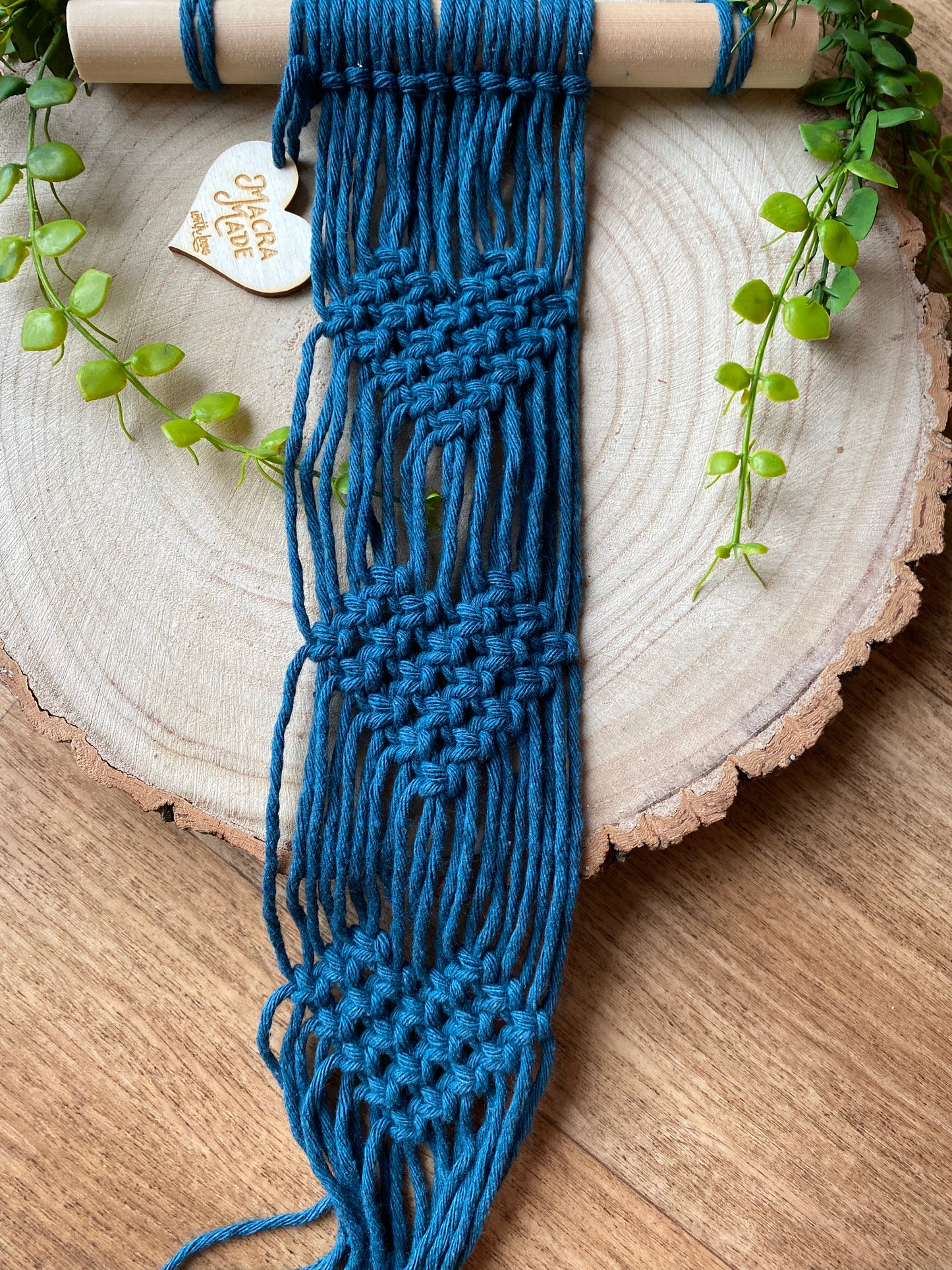 A macrame plant hanger made with petrol blue yarn on a wooden dowel, with knotted heart detail, laid out across a round wooden surface.