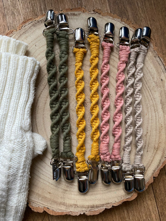 4 pairs of handmade macrame mitten clips lay next to each other on a round wooden surface next to a pair of white mittens. The mitten clips are made with green, yellow, pink and taupe yarn and have metal clips on each end.