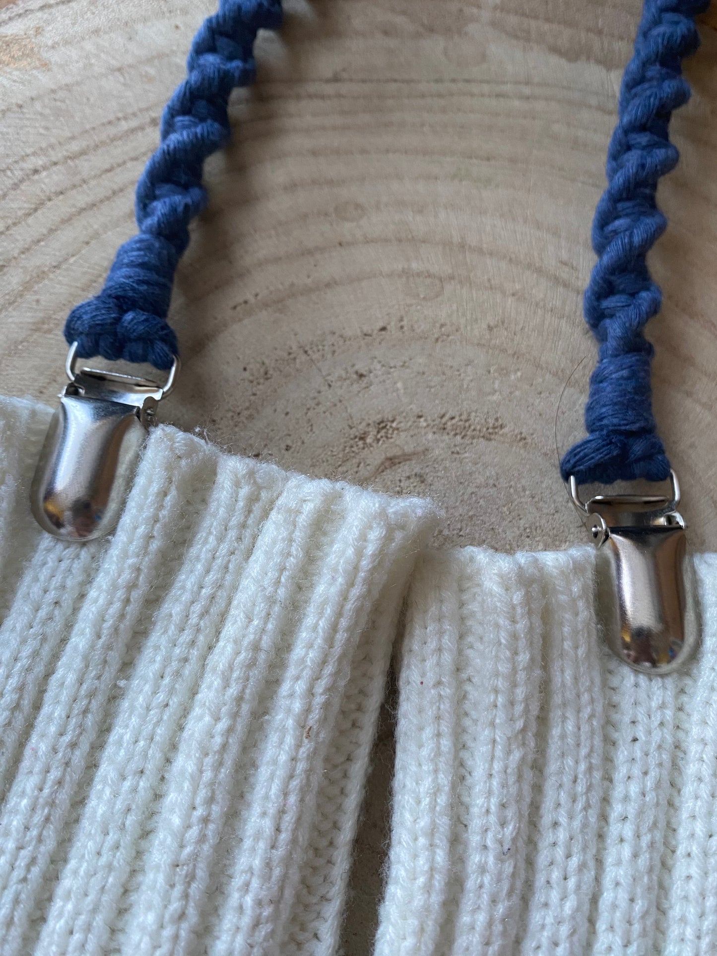 A pair of handmade macrame mitten clips made with blue yarn with metal clips on each end, attached to a pair of white mittens on a wooden board.