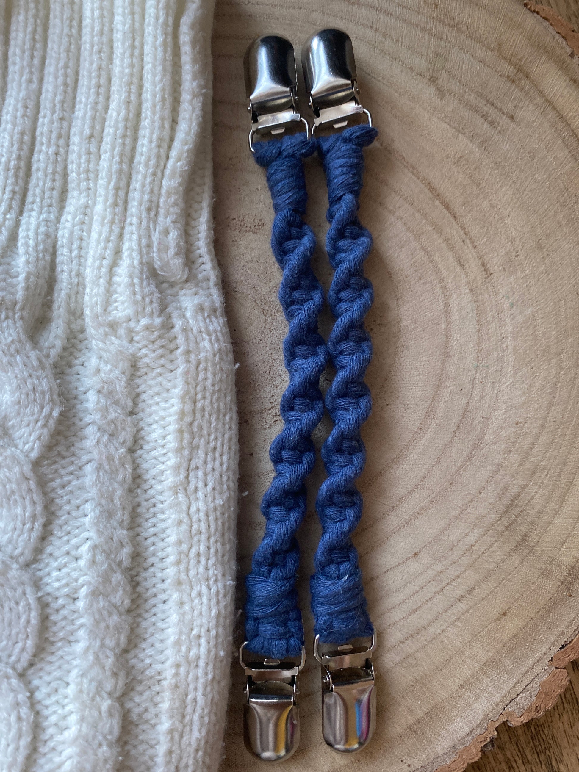 A pair of handmade macrame mitten clips made with blue yarn with metal clips on each end, laid flat next to a pair of white mittens on a wooden board.