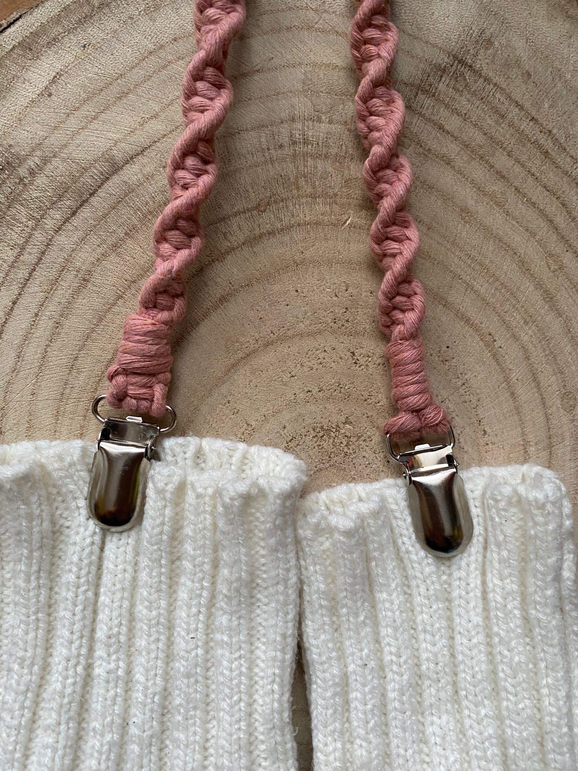 A pair of handmade macrame mitten clips made with blush pink yarn with metal clips on each end, attached to a pair of white mittens on a wooden board.