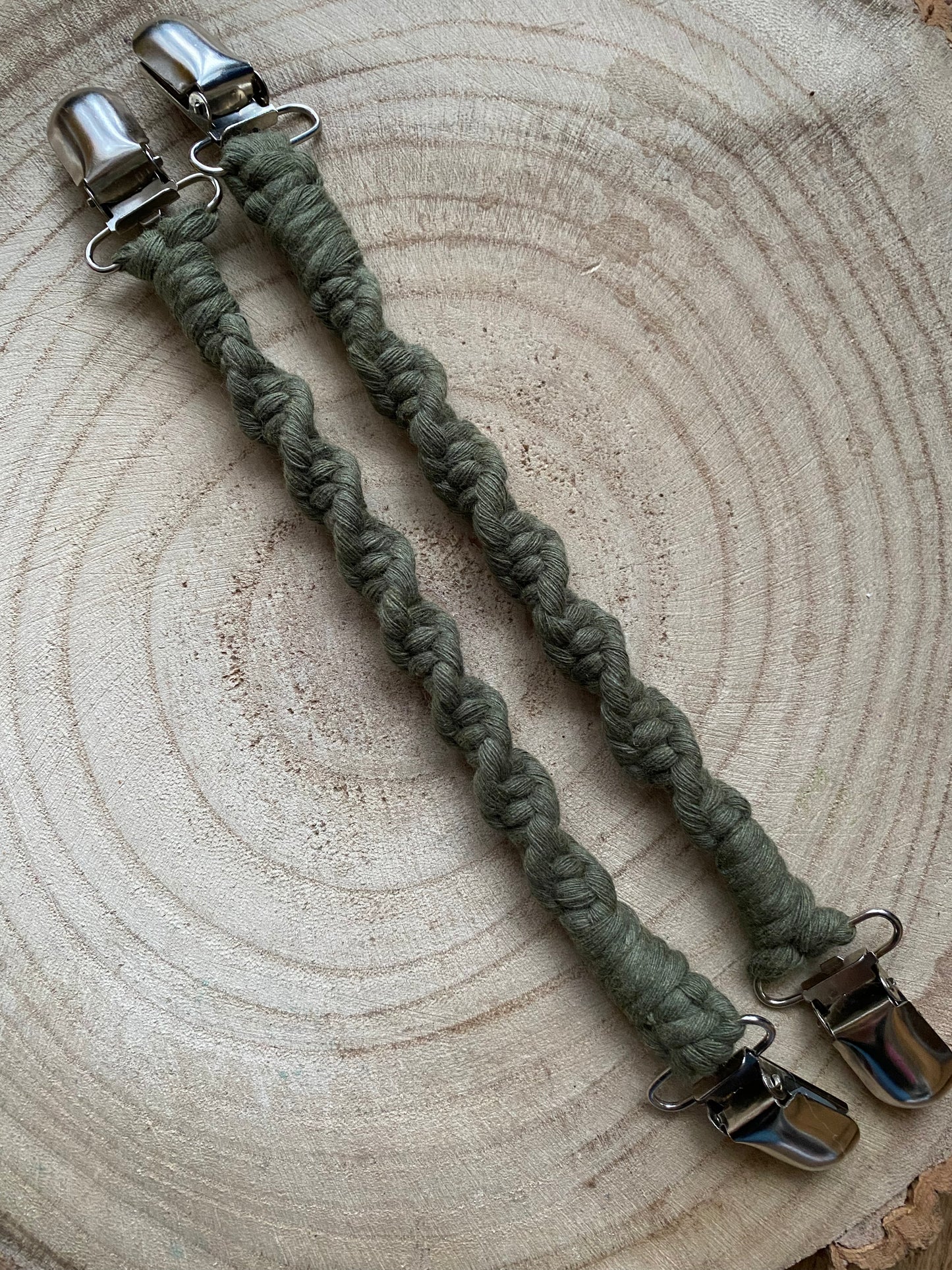 A pair of handmade macrame mitten clips made with avocado green yarn with metal clips on each end, on a wooden board.