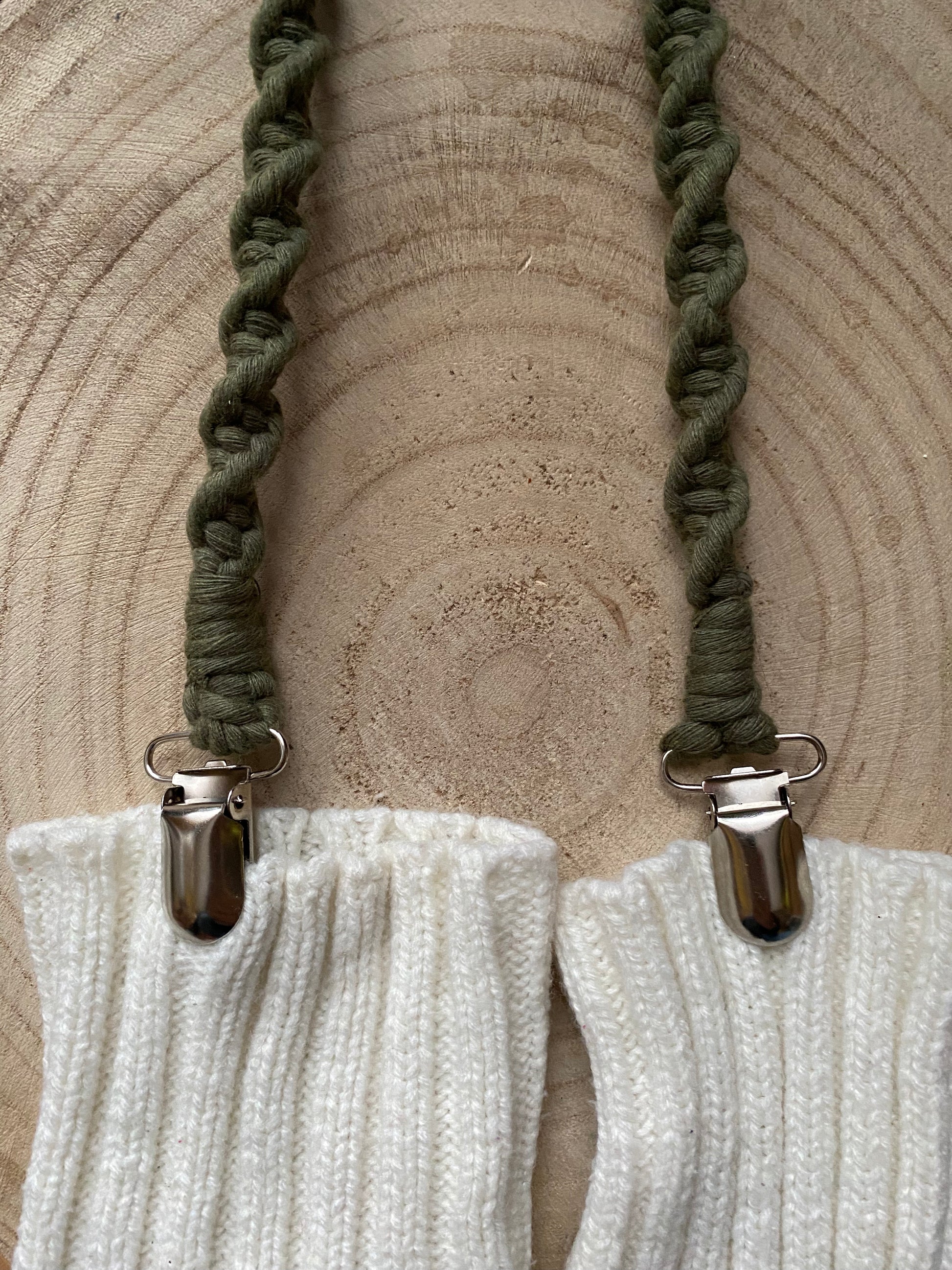 A pair of handmade macrame mitten clips made with green yarn with metal clips on each end, attached to a pair of white mittens on a wooden board.