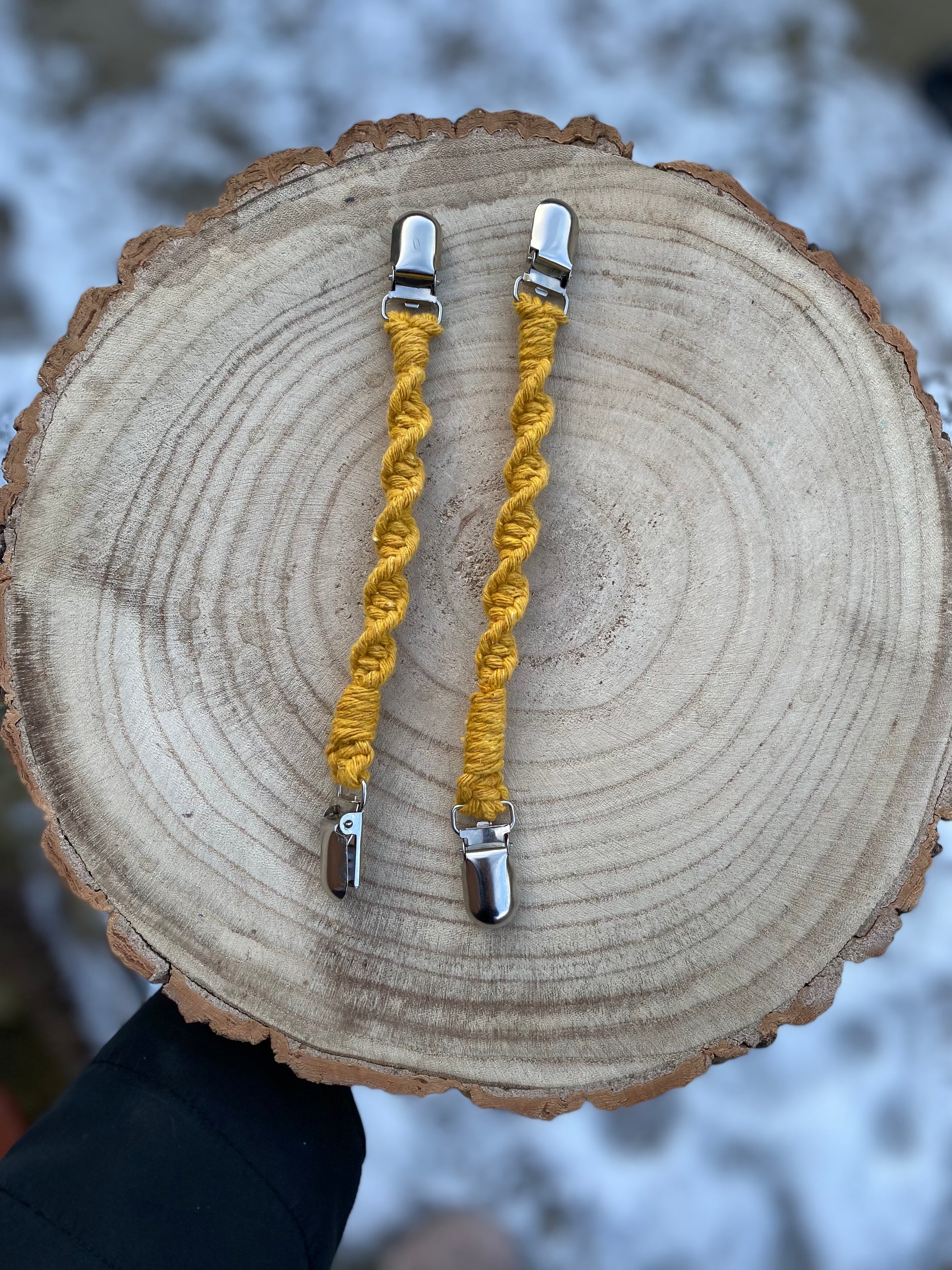 A pair of handmade macrame mitten clips on a round wooden board which is being held outside over snowy ground. The mitten clips are made with mustard yellow yarn and have metal clips on each end.