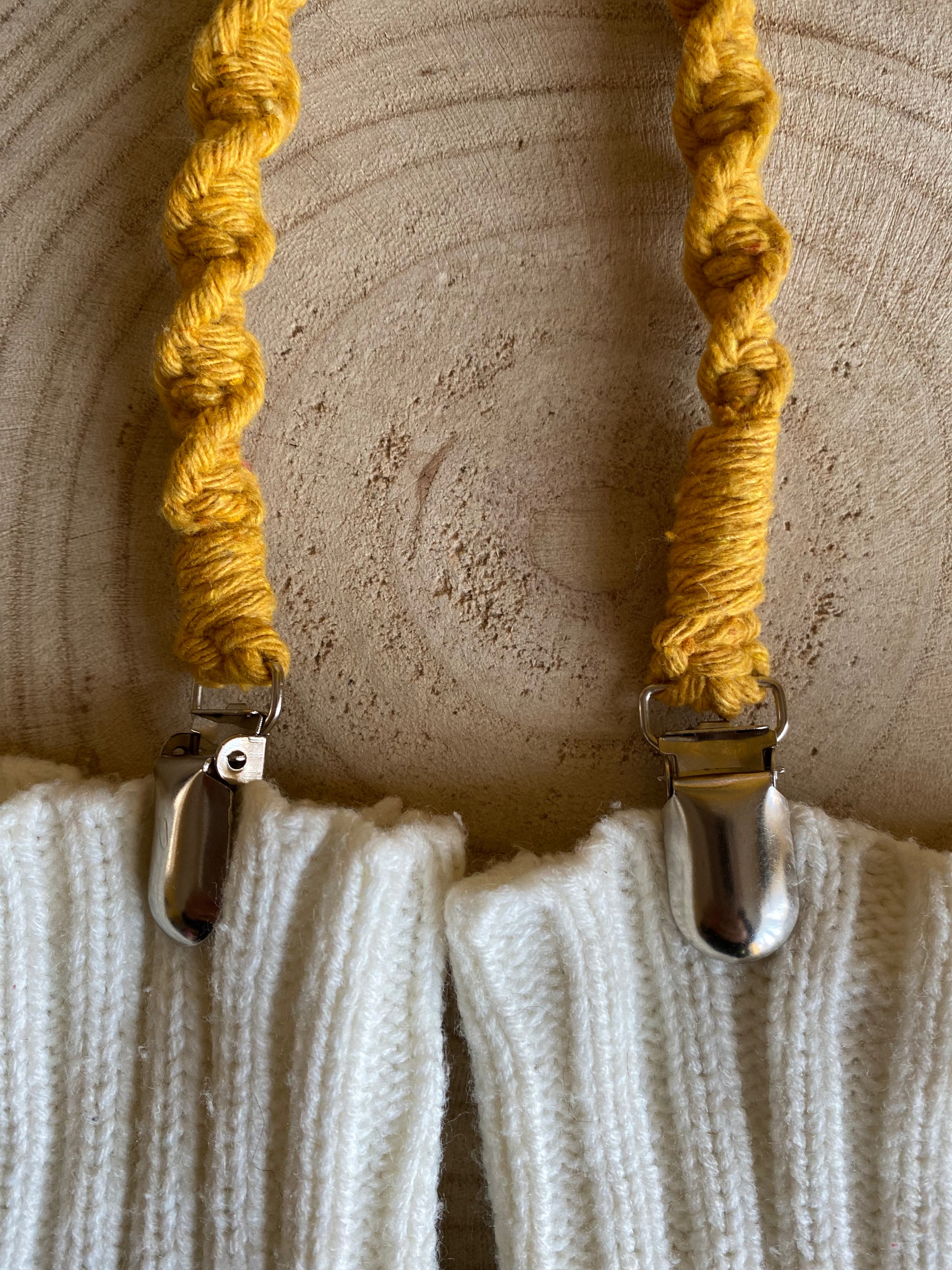 Close up of a pair of handmade macrame mitten clips on a wooden board, attached to a pair of white mittens with metal clips.