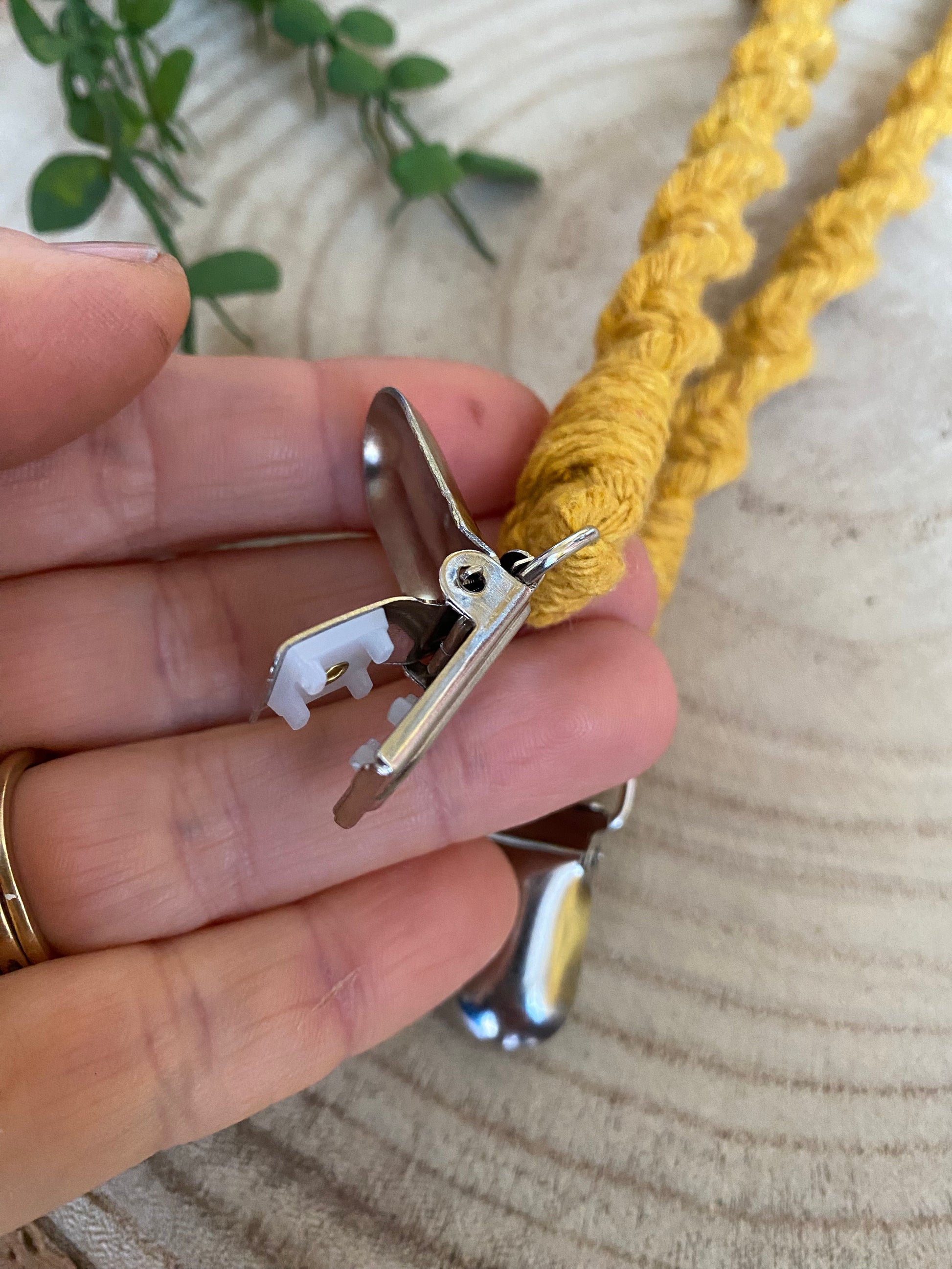 Close up of a hand holding an open metal crocodile clip on the end of a pair of handmade macrame mitten clips made with mustard yellow yarn.