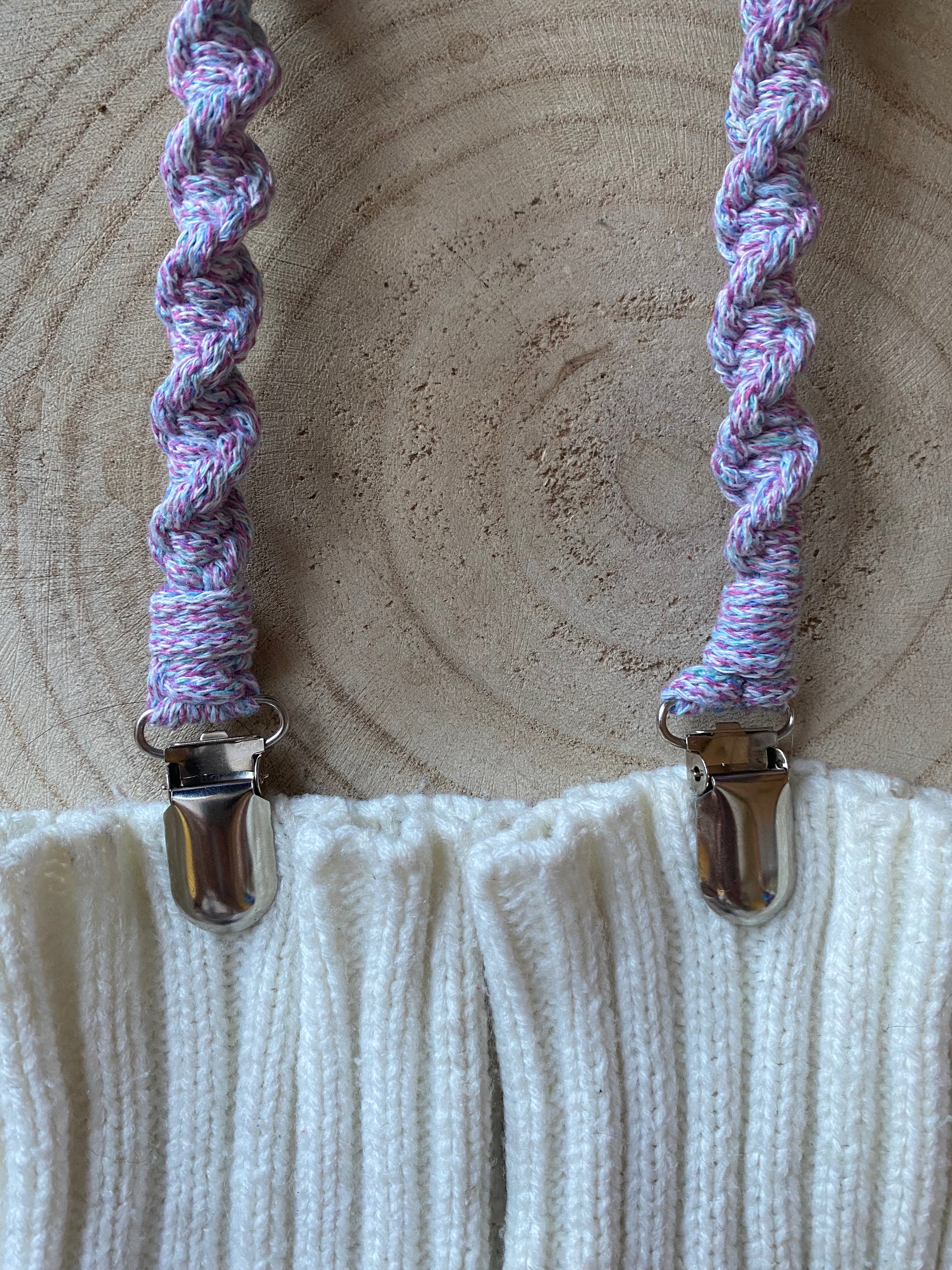 A pair of handmade macrame mitten clips made with pastel yarn with metal clips on each end, attached to a pair of white mittens on a wooden board.