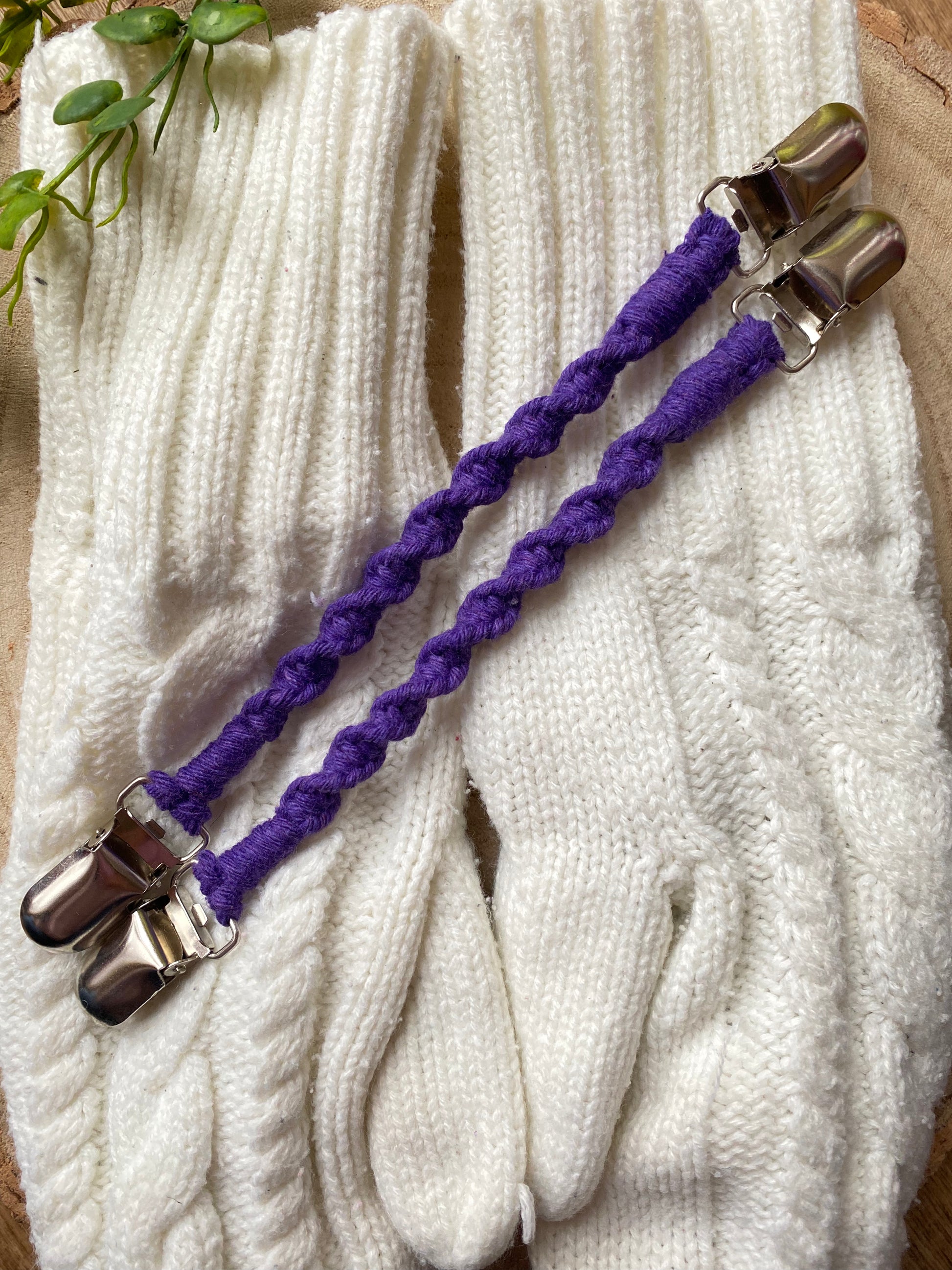 A pair of handmade macrame mitten clips made with purple yarn with metal clips on each end, laid on top of a pair of white mittens on a wooden board.
