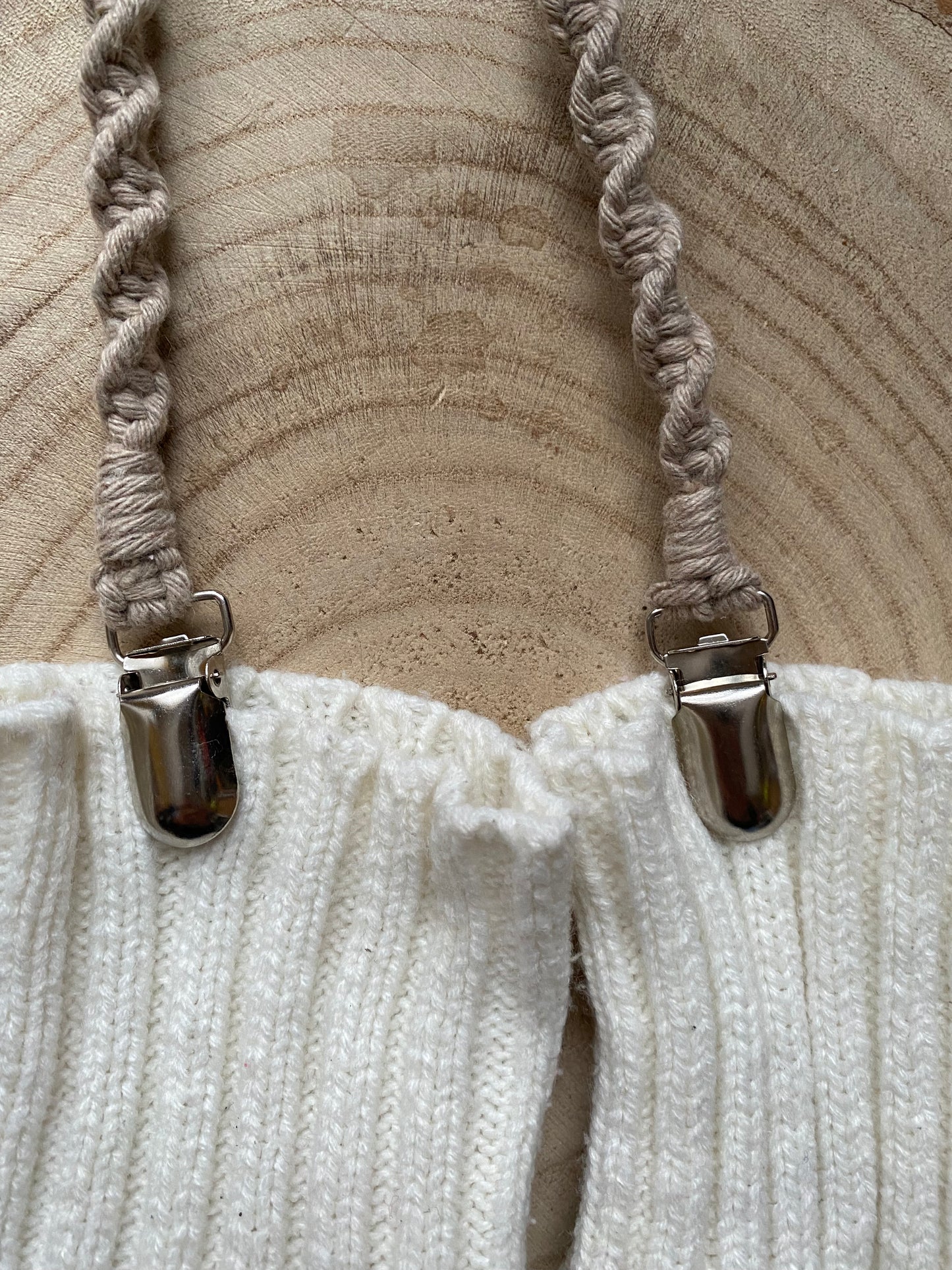 A pair of handmade macrame mitten clips made with taupe yarn with metal clips on each end, attached to a pair of white mittens on a wooden board.
