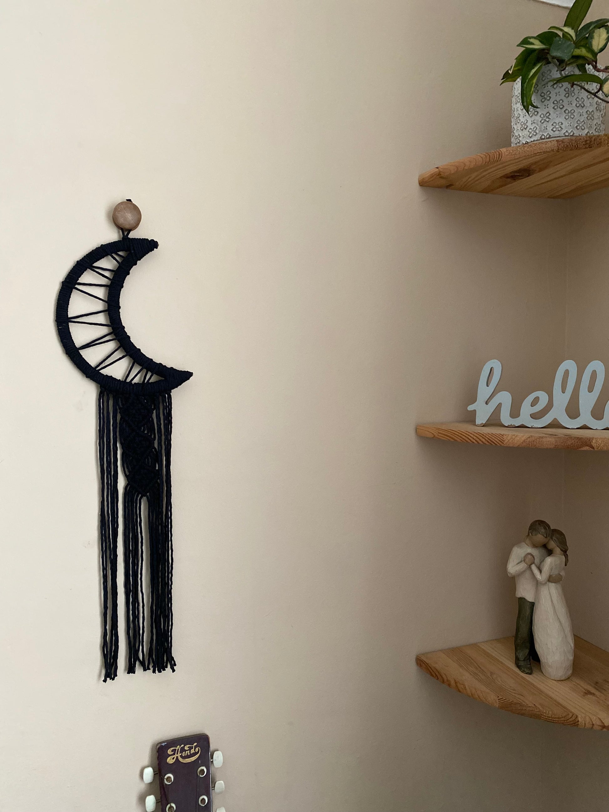 A macrame wall hanging in the shape of a crescent moon, wrapped in black yarn, with long knotted fringe detail hanging on a beige wall next to a set of corner shelves.