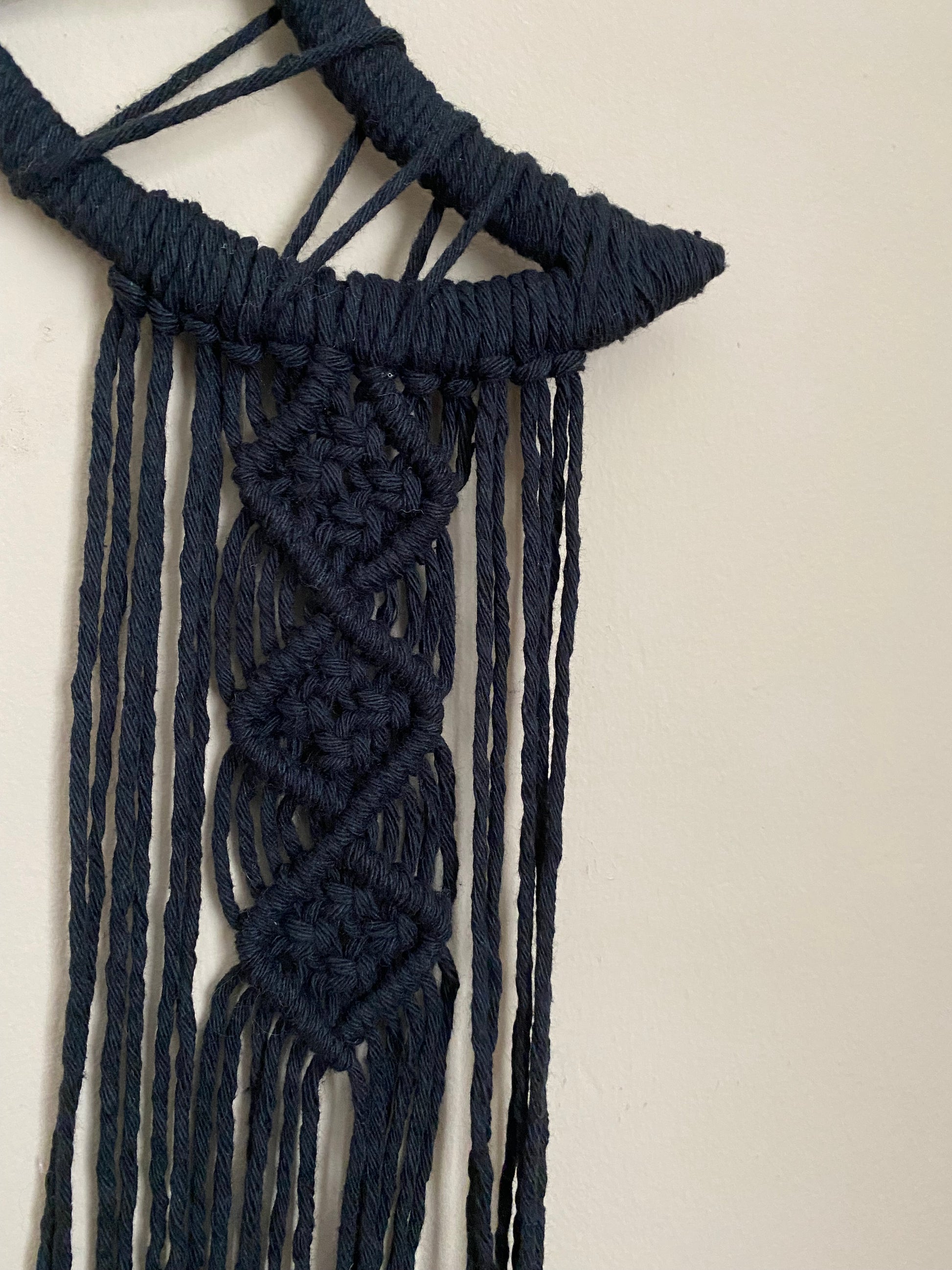 Close up of a macrame wall hanging made with black yarn, with an intricate knot pattern attached to a crescent moon shaped frame.