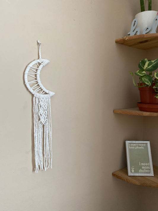 A macrame wall hanging in the shape of a crescent moon, wrapped in natural yarn, with long knotted fringe detail hanging on a beige wall next to a set of wooden corner shelves.