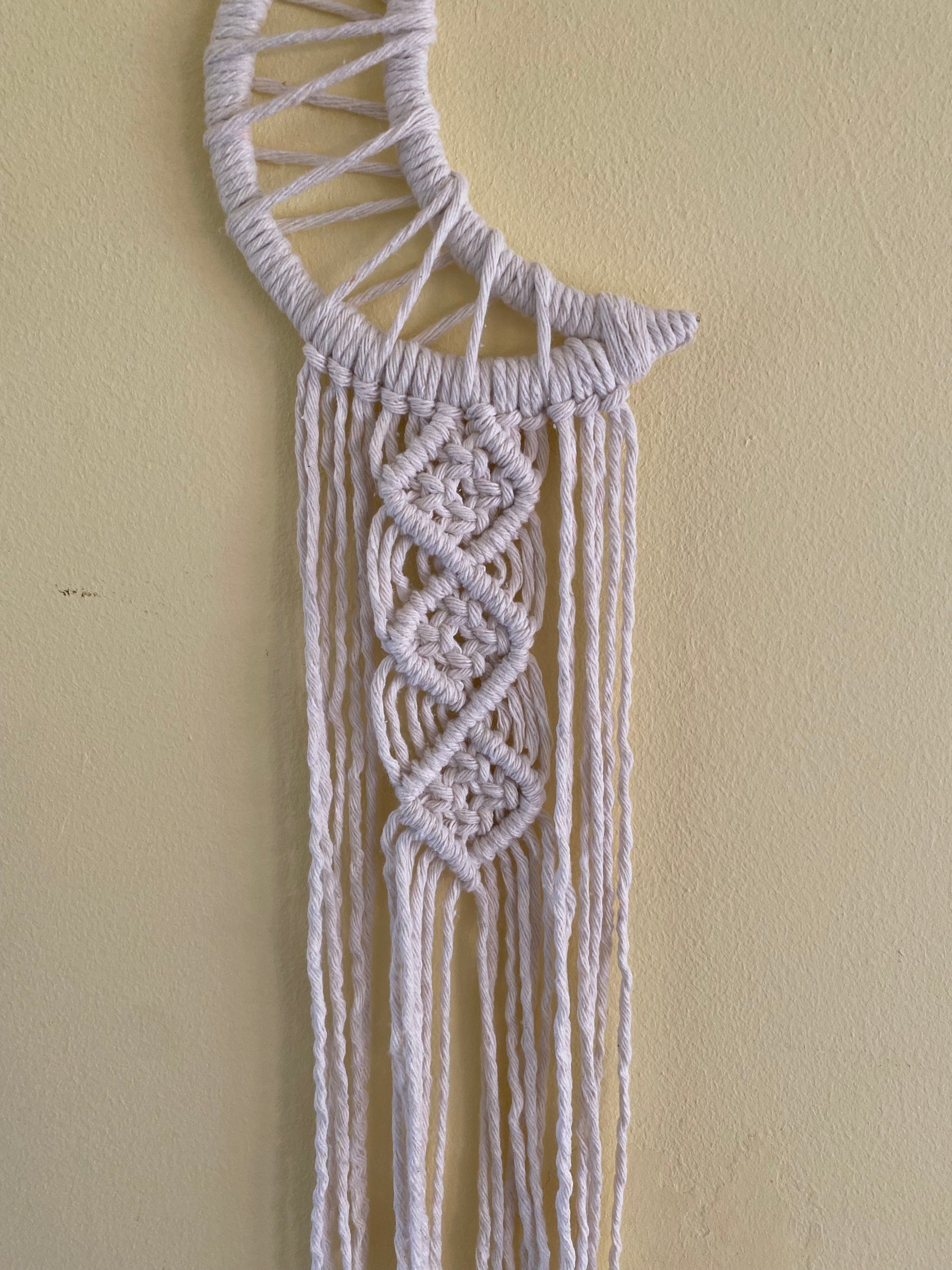 Close up of a macrame wall hanging in the shape of a crescent moon, wrapped in natural yarn, with knotted fringe detail.