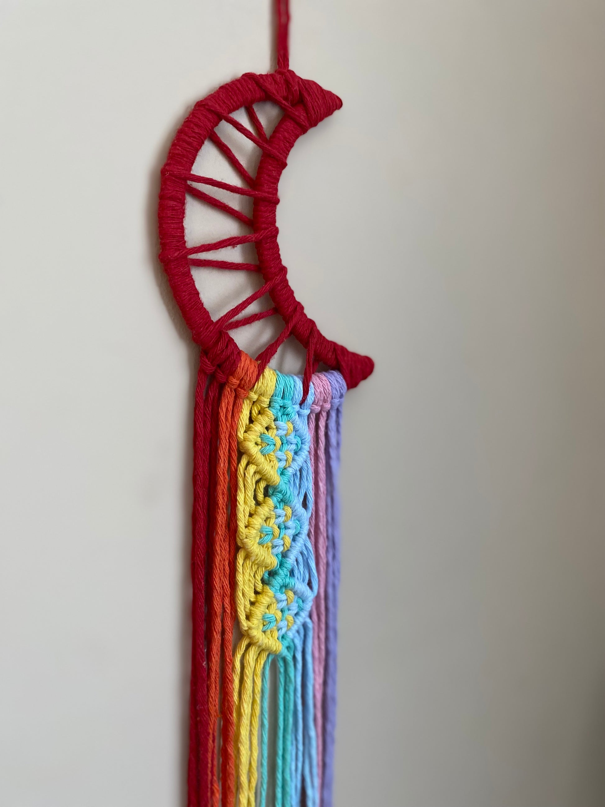 A close up of a macrame wall hanging in the shape of a crescent moon, wrapped in red yarn, with rainbow knotted fringe detail hanging on a beige wall.