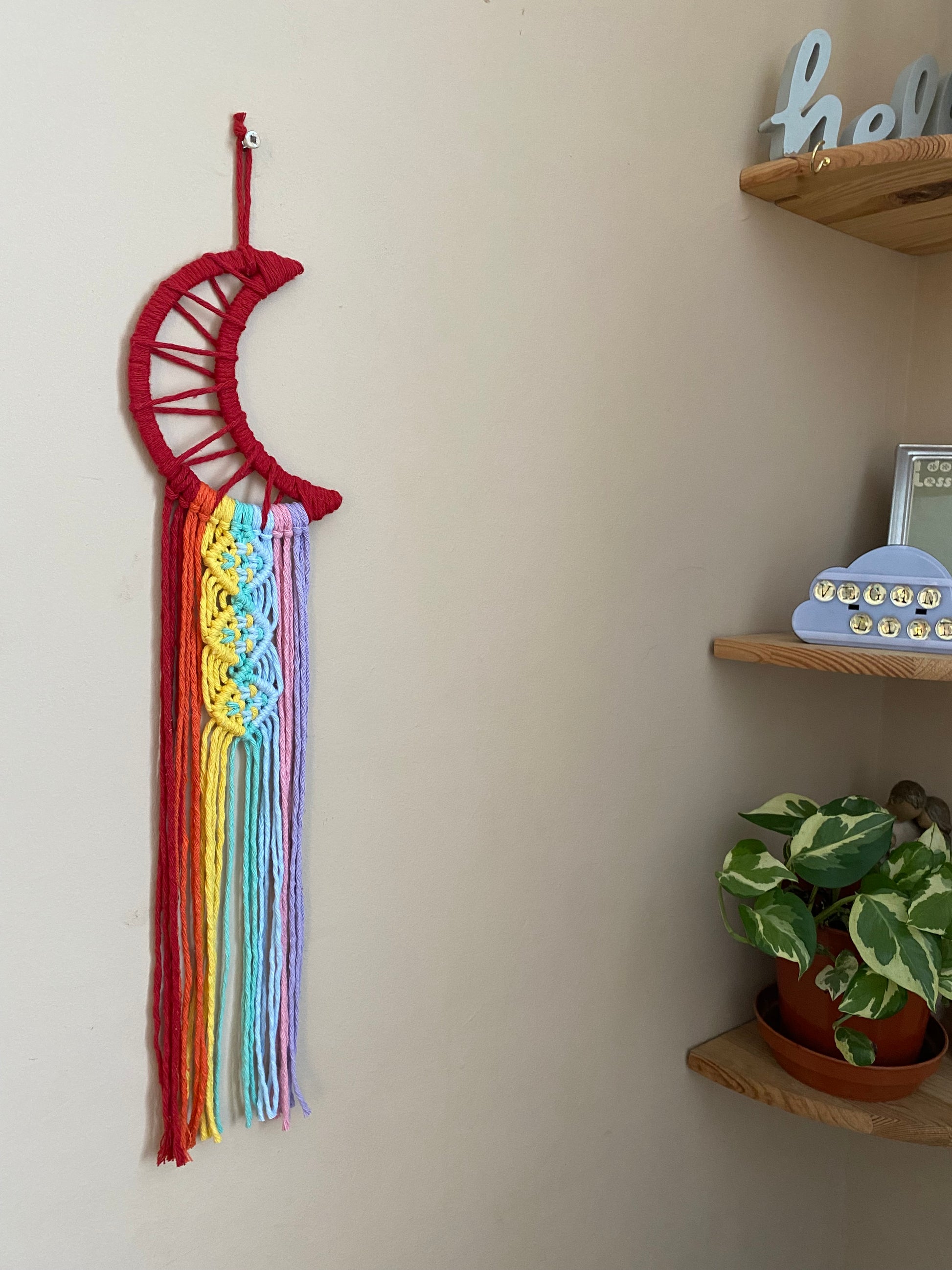 A macrame wall hanging in the shape of a crescent moon, wrapped in red yarn, with long rainbow knotted fringe detail hanging on a beige wall next to a set of corner shelves.