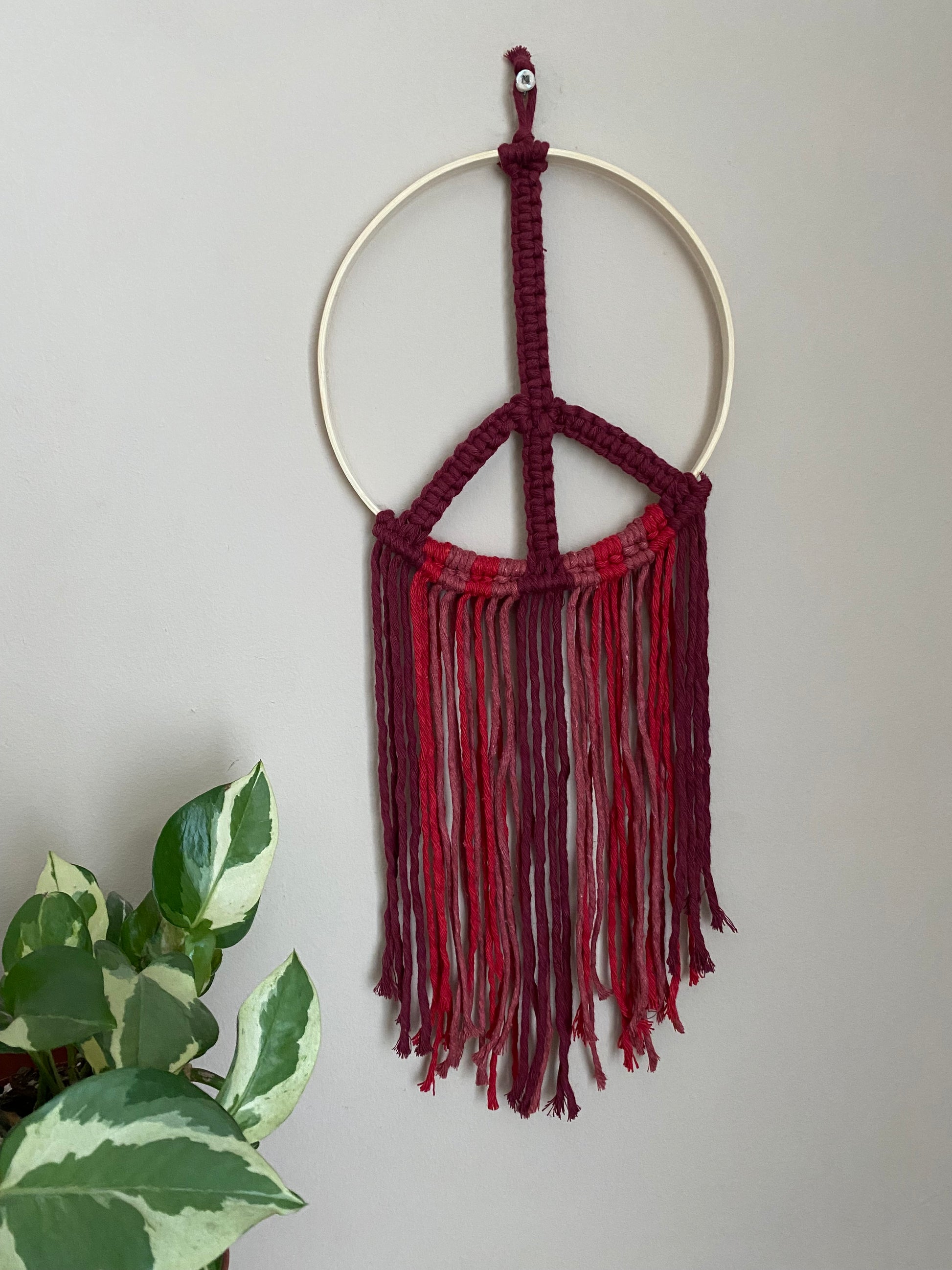 Macrame peace sign wall hanging decoration, knotted with red cotton yarns on to a round bamboo hoop, hanging against a beige wall next to a green houseplant.