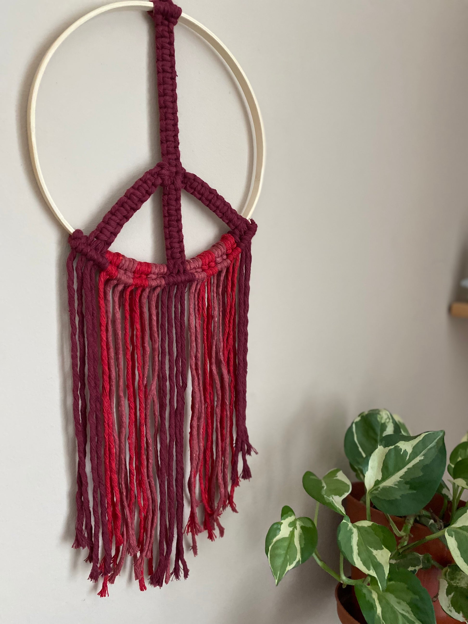Close up side angle of a macrame peace sign wall hanging decoration, knotted with red cotton yarns on to a round bamboo hoop, hanging against a beige wall next to a green houseplant.