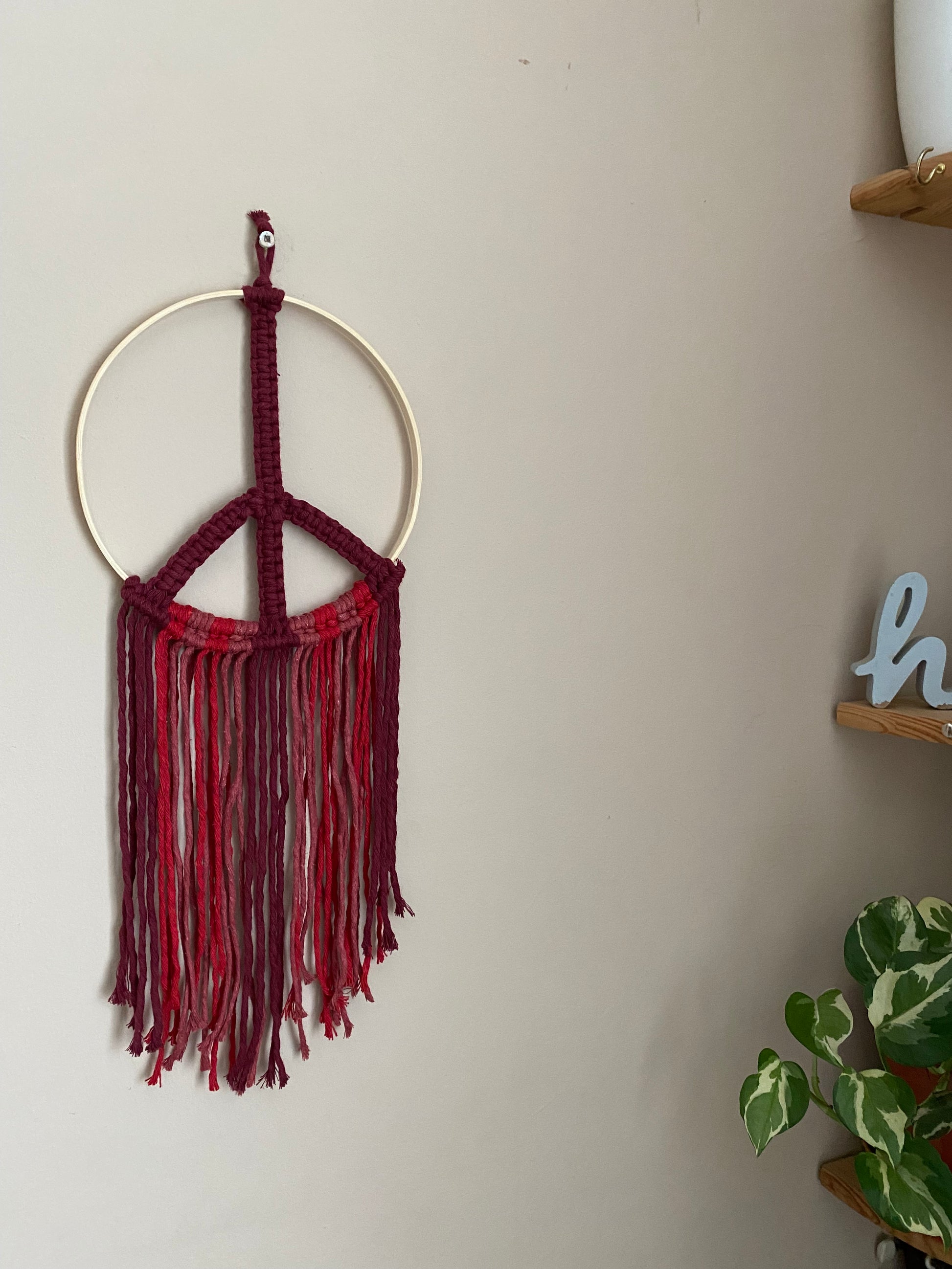 Macrame peace sign wall hanging decoration, knotted with red cotton yarns on to a round bamboo hoop, hanging against a beige wall next to a green houseplant and a set of corner shelves.