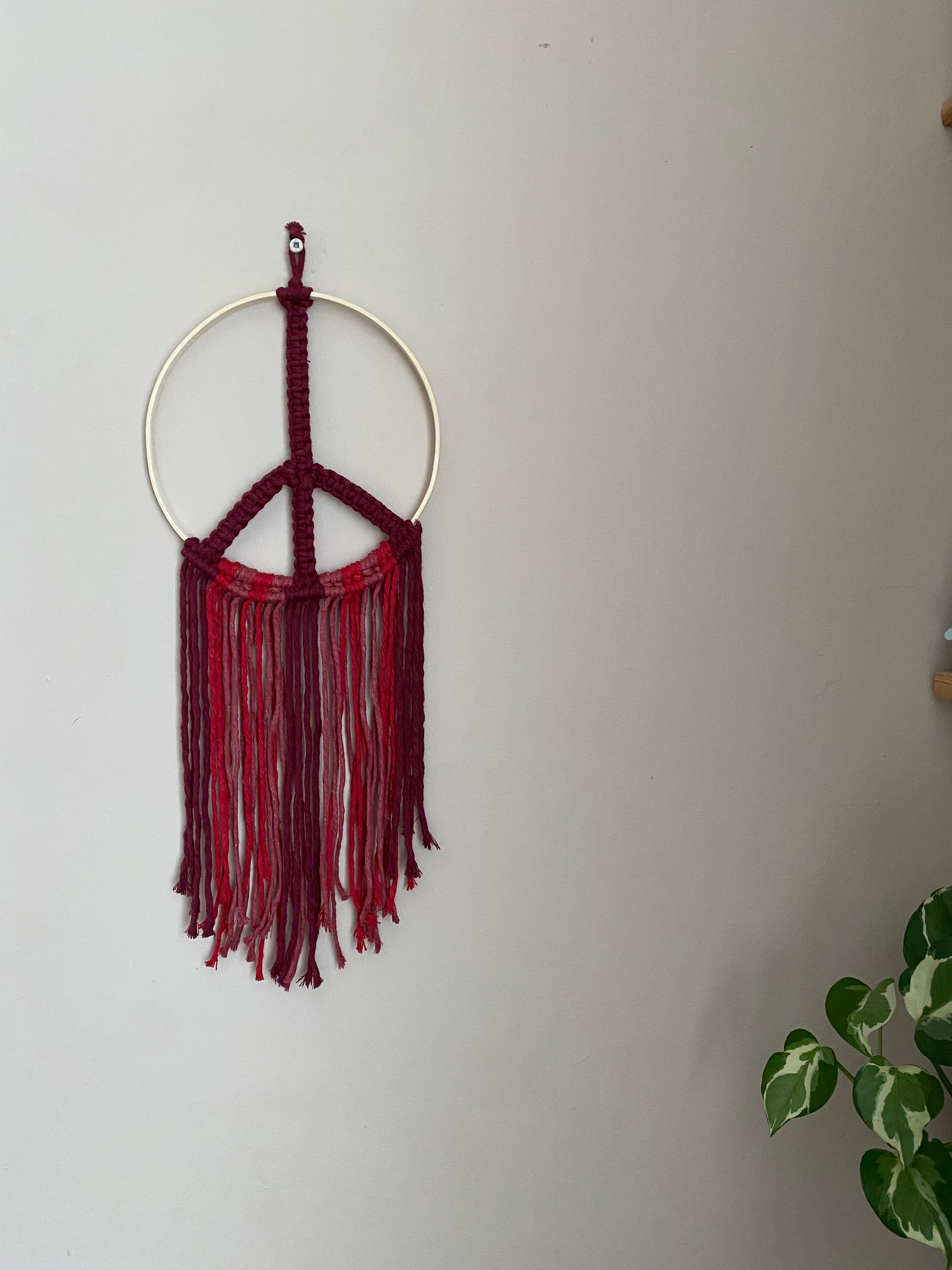 Macrame peace sign wall hanging decoration, knotted with red cotton yarns on to a round bamboo hoop, hanging against a beige wall next to a green houseplant.