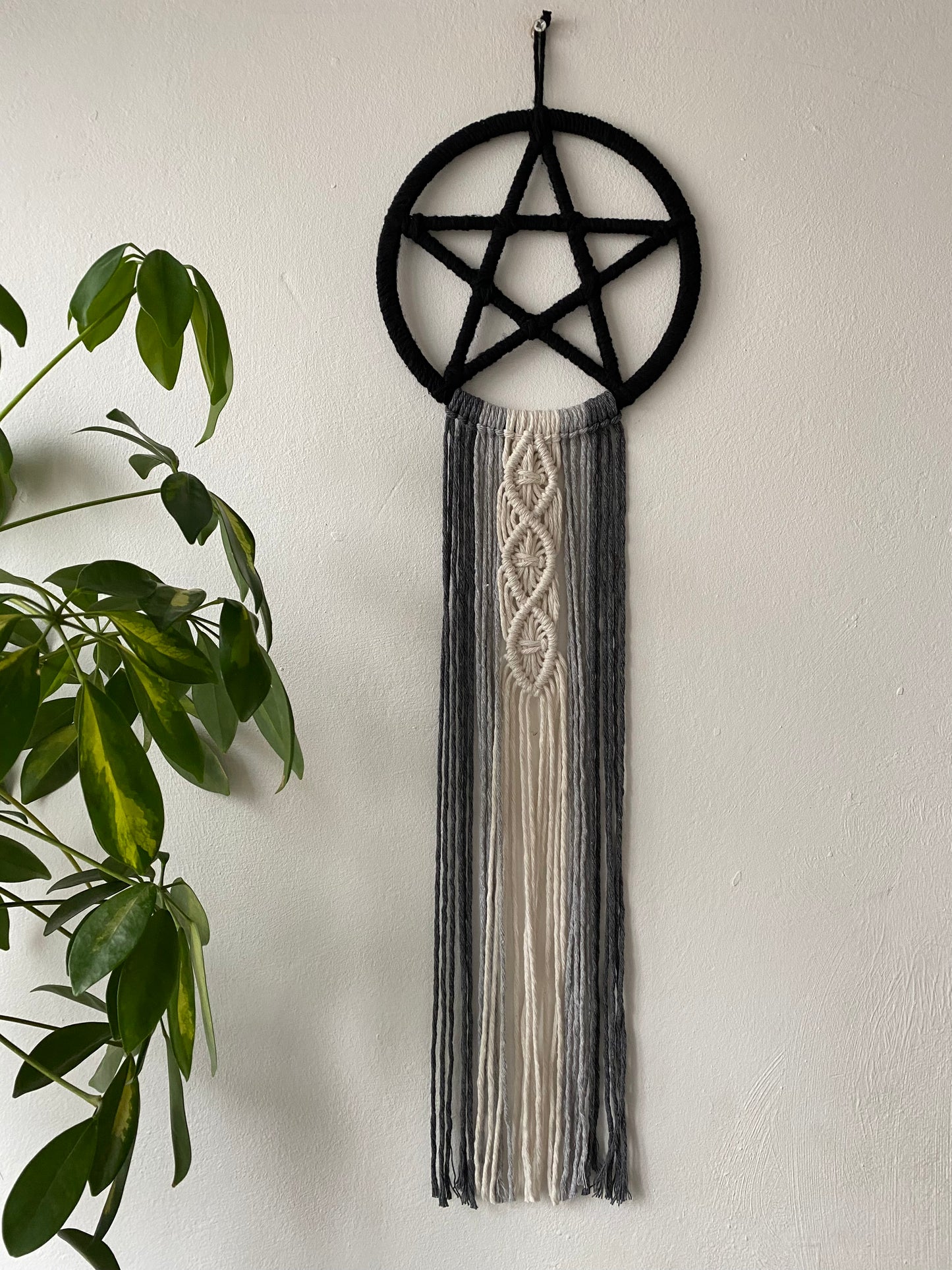 Handmade macrame pentagram wall hanging made with black, grey and natural yarns with long flowing tails, hanging against a white wall next to a tall green houseplant.