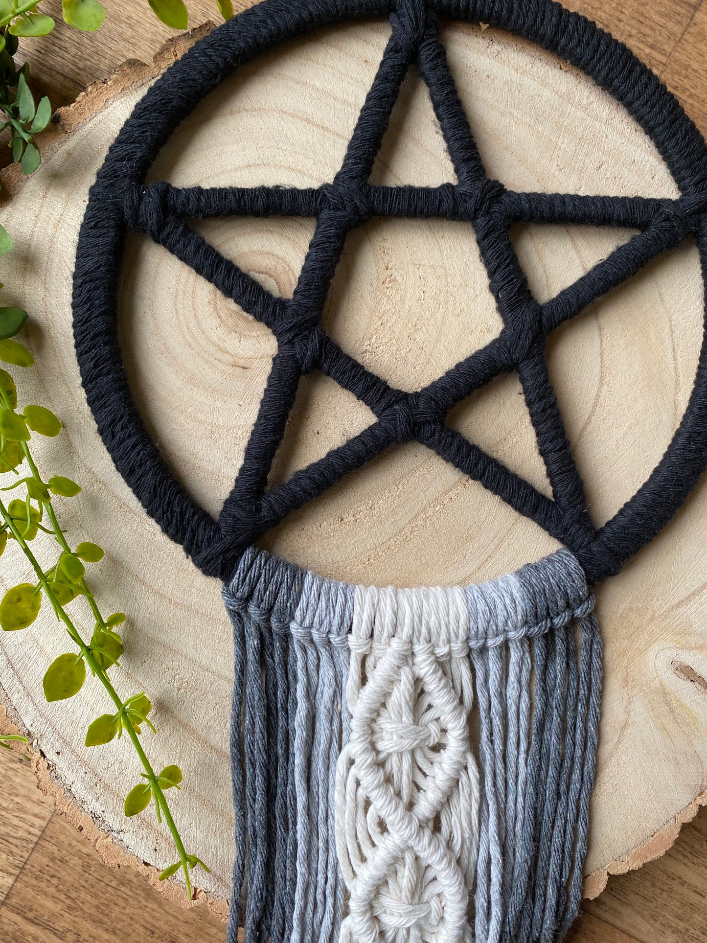 Handmade macrame pentagram wall hanging made with black, grey and natural yarns laid across a wooden board.