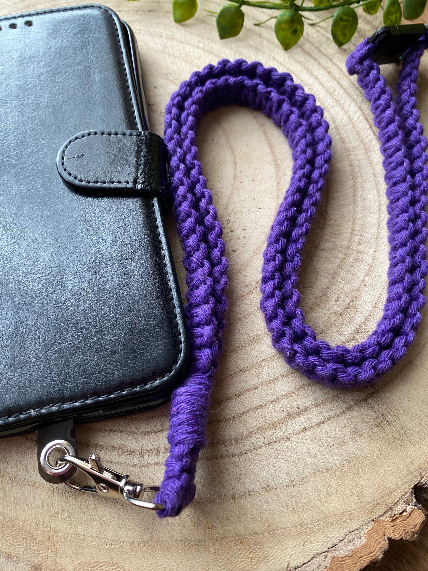 A black leather phone case on a wooden board with a macrame phone lanyard made with purple yarn attached to it with a metal clasp.