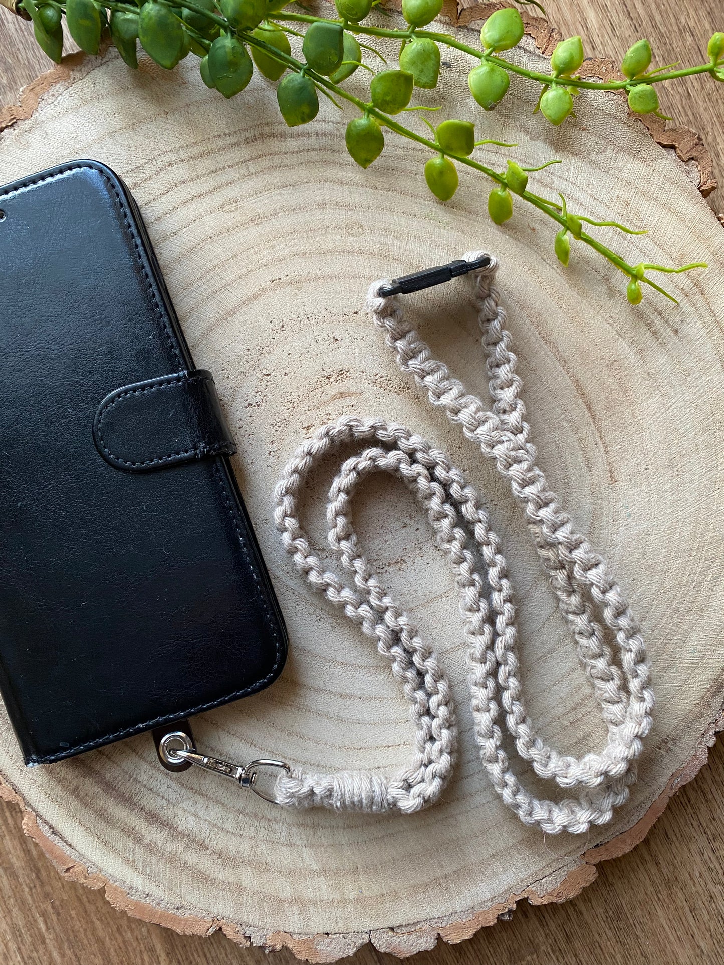 A black leather phone case on a wooden board with a macrame phone lanyard made with taupe yarn attached to it with a metal clasp.