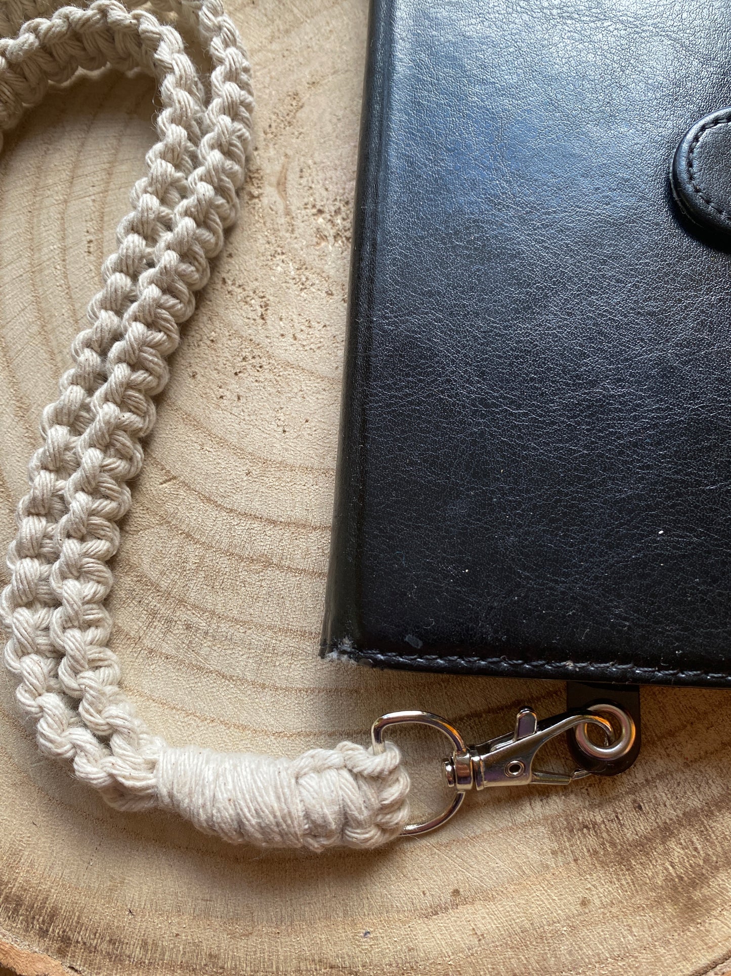 Close up of a black leather phone case on a wooden board with a macrame phone lanyard made with taupe yarn attached to it with a metal clasp.