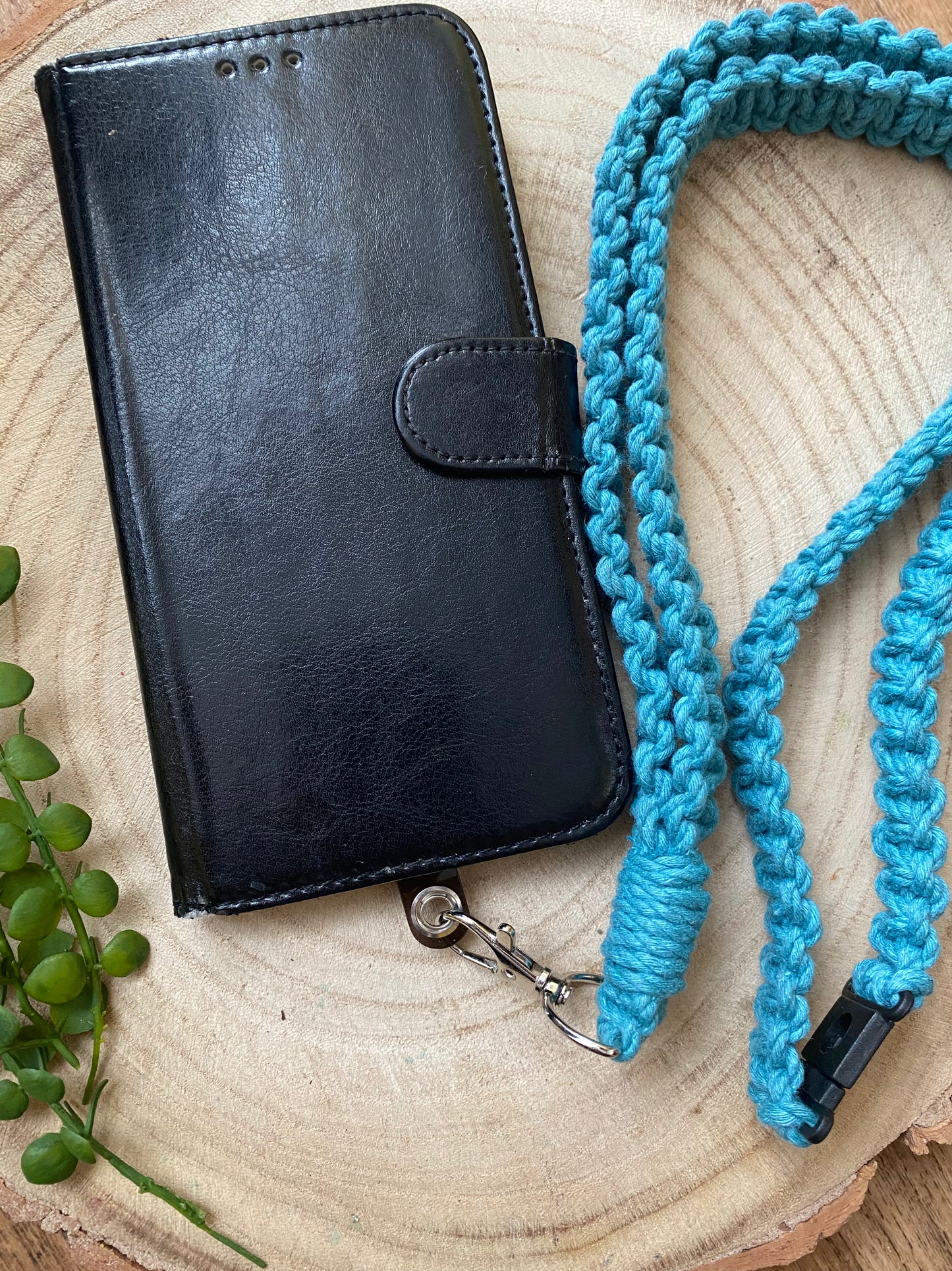 A black leather phone case on a wooden board with a teal blue macrame phone lanyard attached to it with a metal clasp.