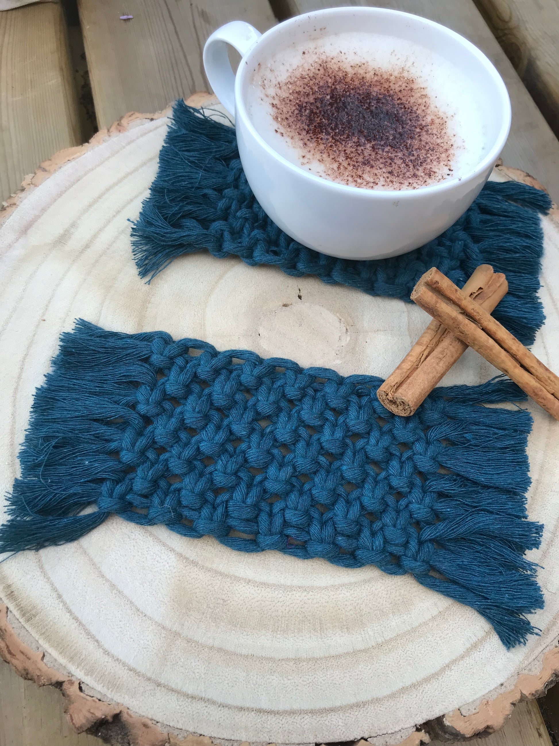 A set of macrame rectangular coasters made with blue, soft yarn, with knotted detail and fringed edges, is placed on a circular wooden board. One coaster has a white mug with coffee on top of it and there are two cinnamon sticks next to the cup for decoration.