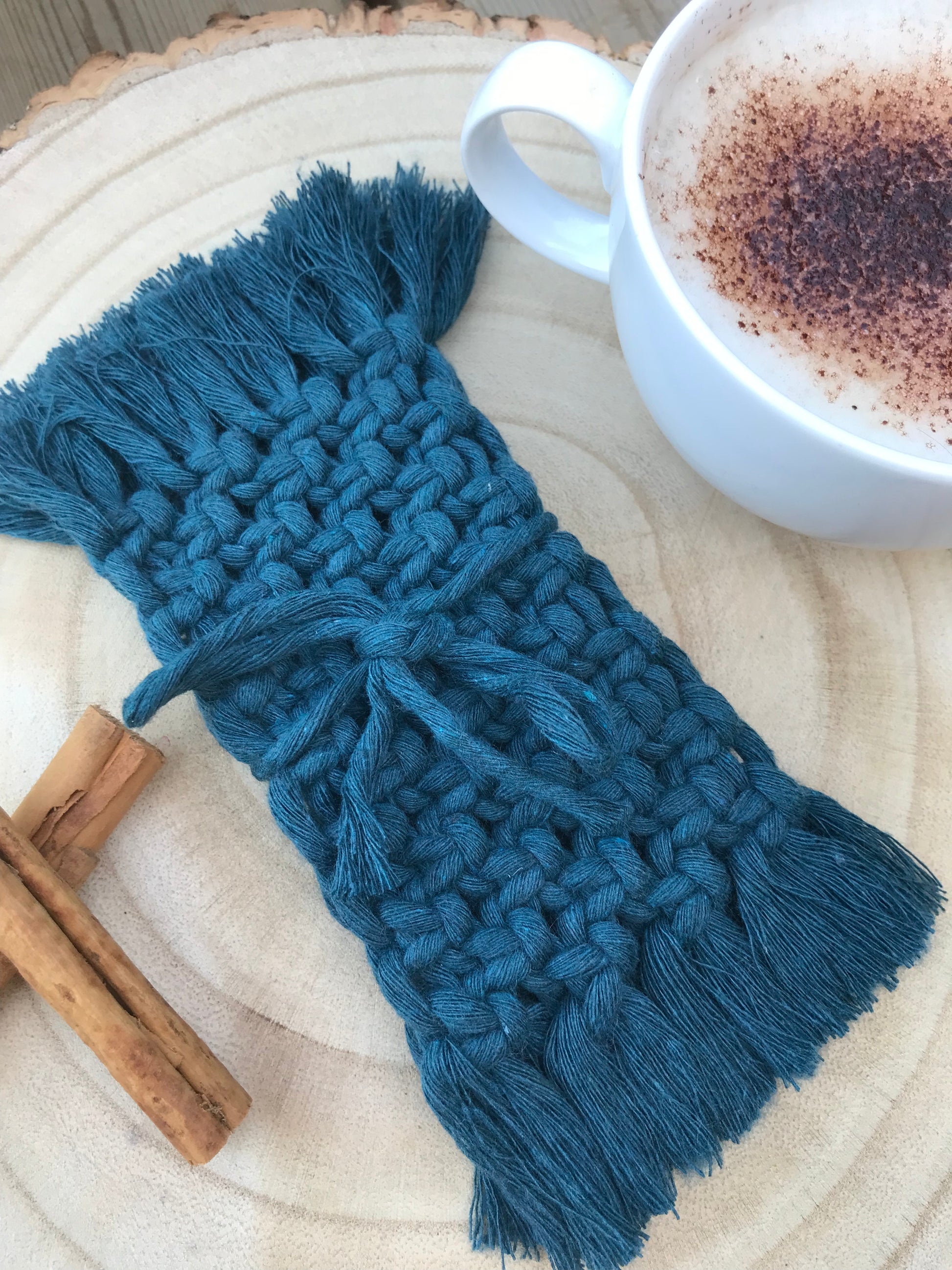 A set of macrame rectangular coasters made with blue yarn, with knotted detail and fringed edges, is placed on a circular wooden board, tied together with a piece of matching blue yarn. There is a white mug with coffee in it, and two cinnamon sticks next to the coasters for decoration.