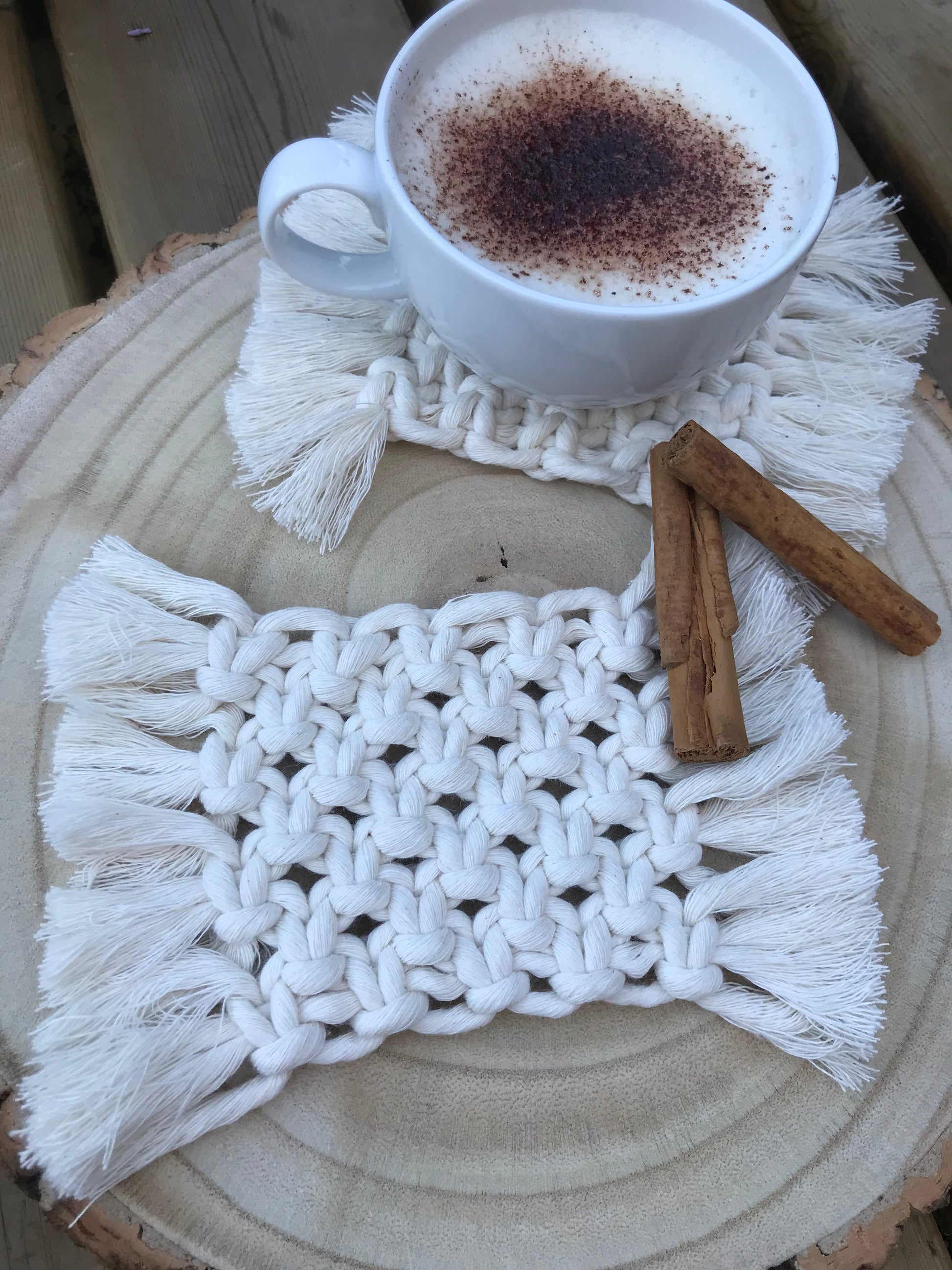 A set of macrame rectangular coasters made with natural coloured, soft yarn, with knotted detail and fringed edges, is placed on a circular wooden board. One coaster has a white mug with coffee on top of it and there are two cinnamon sticks next to the cup for decoration.