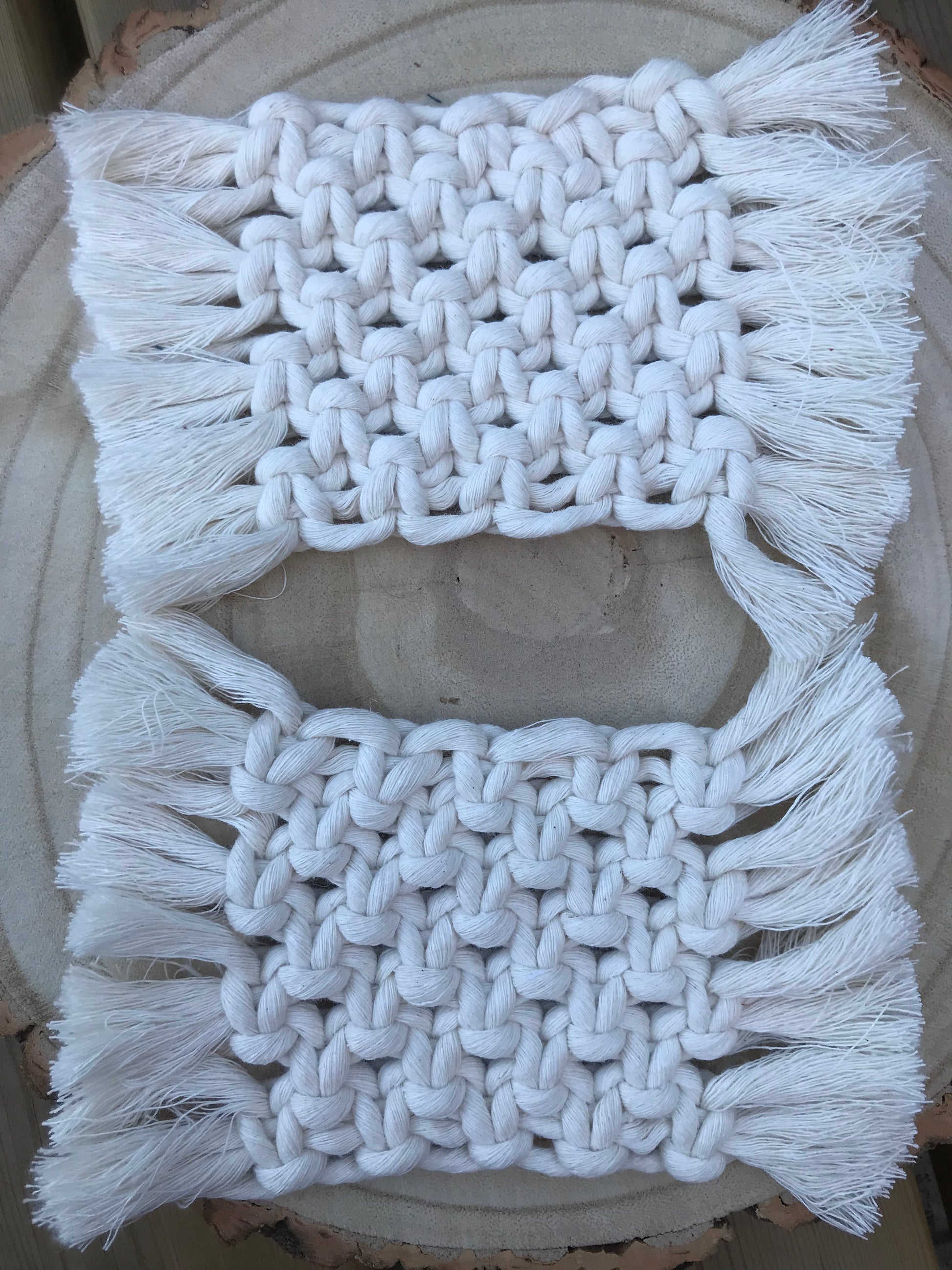 A set of macrame rectangular coasters made with natural coloured, soft yarn, with knotted detail and fringed edges, is placed on a circular wooden board.