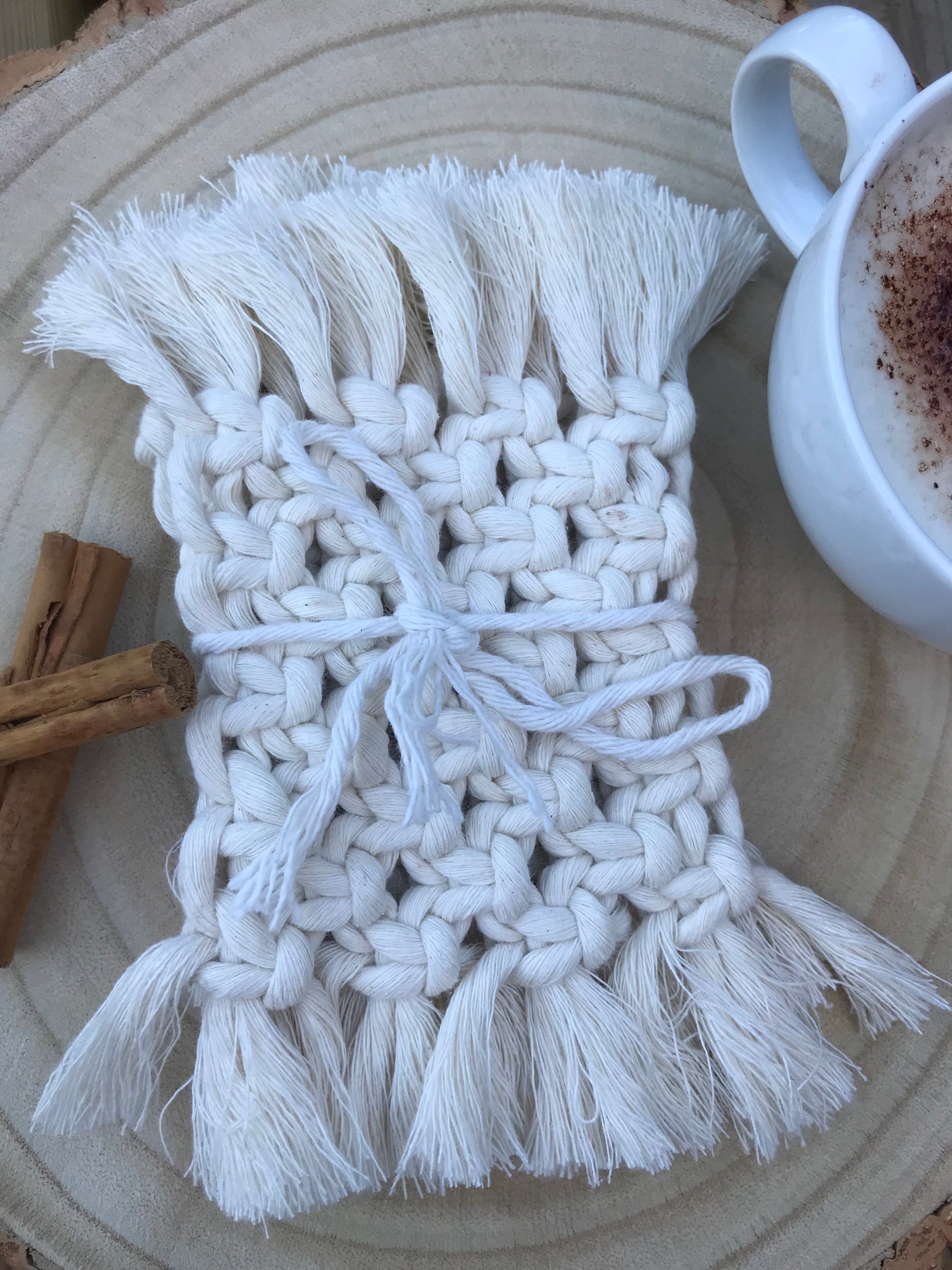 A set of macrame rectangular coasters made with natural coloured, soft yarn, with knotted detail and fringed edges, is placed on a circular wooden board, tied together with a matching yarn bow. There is a white mug with coffee in it, and two cinnamon sticks next to the coasters for decoration.