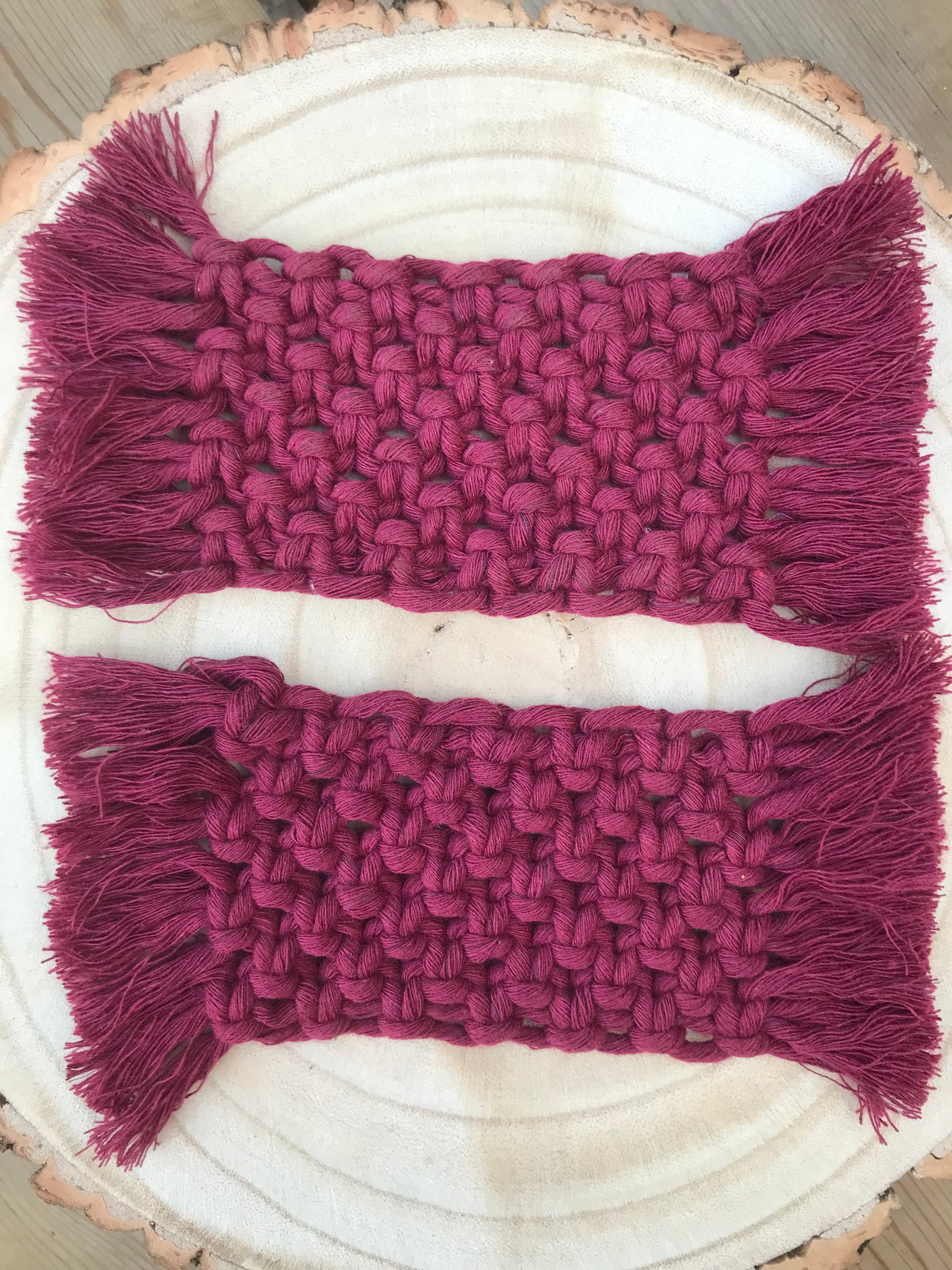 A set of macrame rectangular coasters made with deep red coloured, soft yarn, with knotted detail and fringed edges, is placed on a circular wooden board.