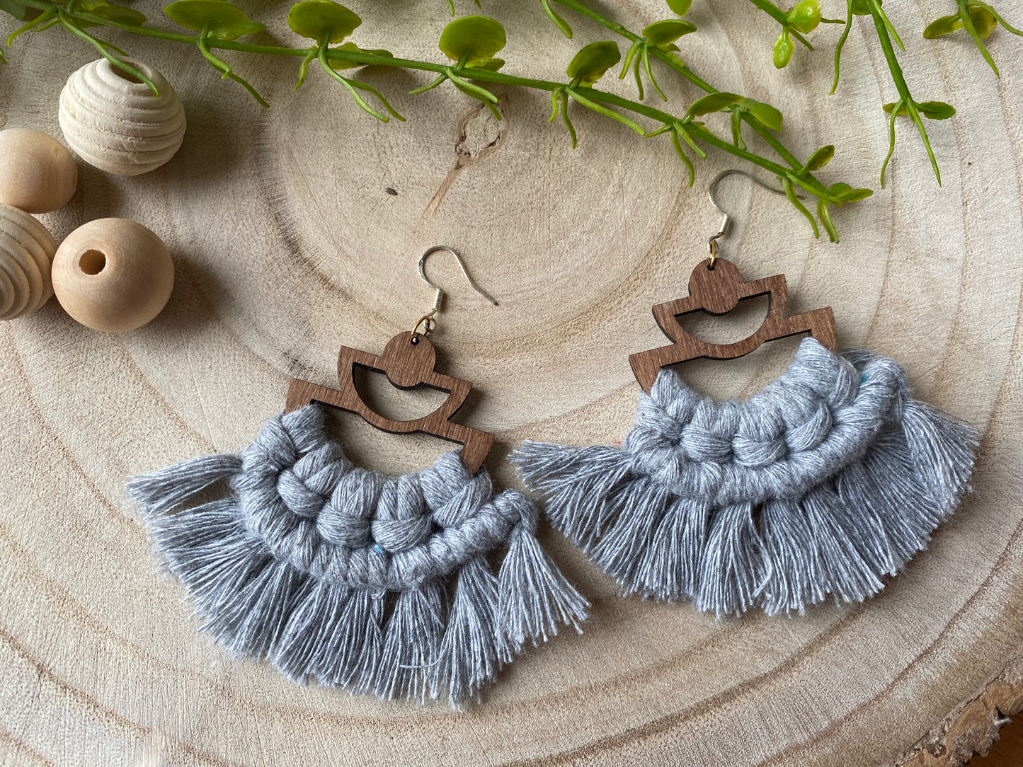 A pair of handmade macrame earrings made with grey yarn, on wooden semi circle findings with silver hooks, and neat fringed detail, placed on a wooden surface surrounded by beads and greenery.