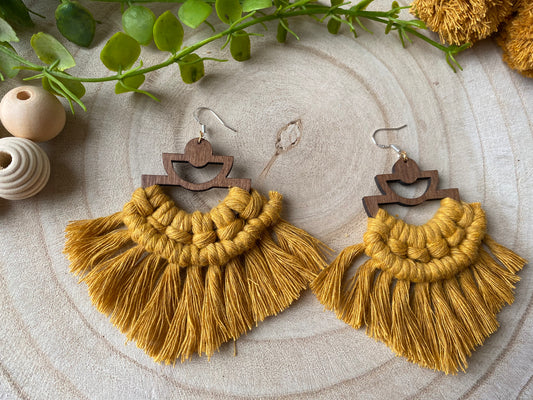 A pair of handmade macrame earrings made with mustard yellow yarn, on wooden semi circle findings with silver hooks, and neat fringed detail, placed on a wooden surface surrounded by beads and greenery.