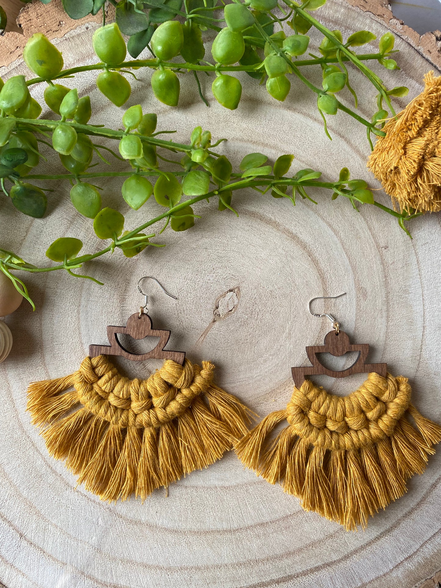 A pair of handmade macrame earrings made with mustard yellow yarn, on wooden semi circle findings with silver hooks, and neat fringed detail, placed on a wooden surface surrounded by beads and greenery.