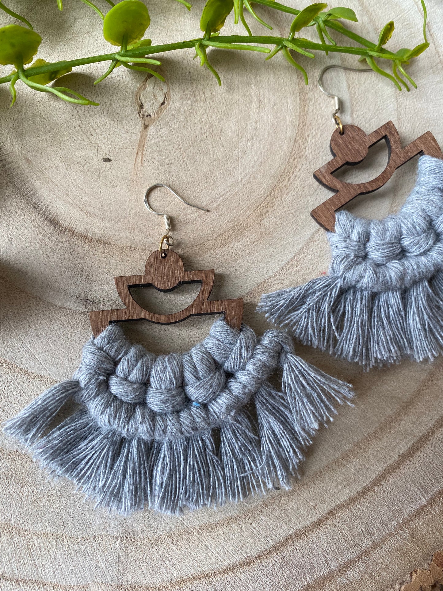 A pair of handmade macrame earrings made with grey yarn, on wooden semi circle findings with silver hooks, and neat fringed detail, placed on a wooden surface surrounded by greenery.