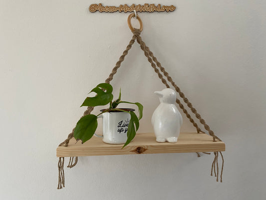 Handmade macramé hanging shelf with a single wooden base, suspended from a knotted recycled cotton cord in a beige cream colour. The boho-style hanger features decorative macramé knots and loops, with a smooth, rectangular wooden shelf at the bottom and a wooden hanging ring at the top. On the shelf is a small plant and a white penguin ornament.