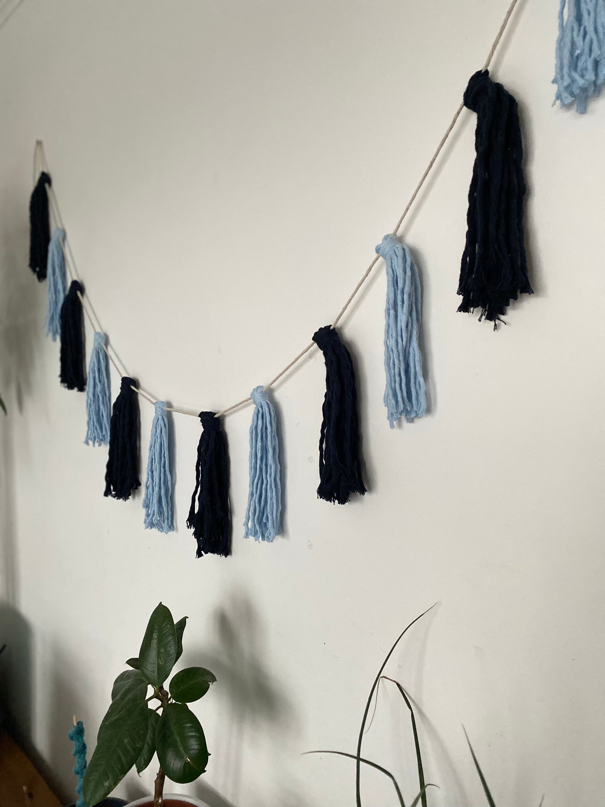 Macrame tassel garland, in shades of blue, hanging on a white wall. There is a small green plant nearby.