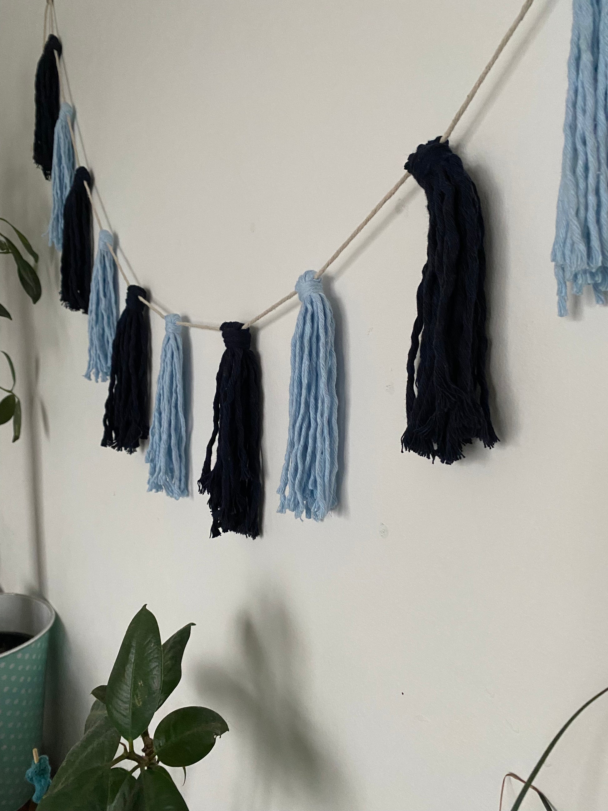 Macrame tassel garland, in shades of blue, hanging on a white wall. There is a small green plant nearby.